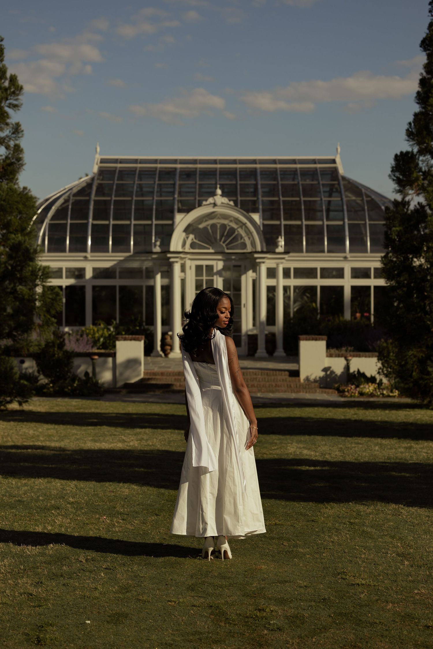 Woman in white dress stands before elegant glass greenhouse conservatory in lush green garden at golden hour.