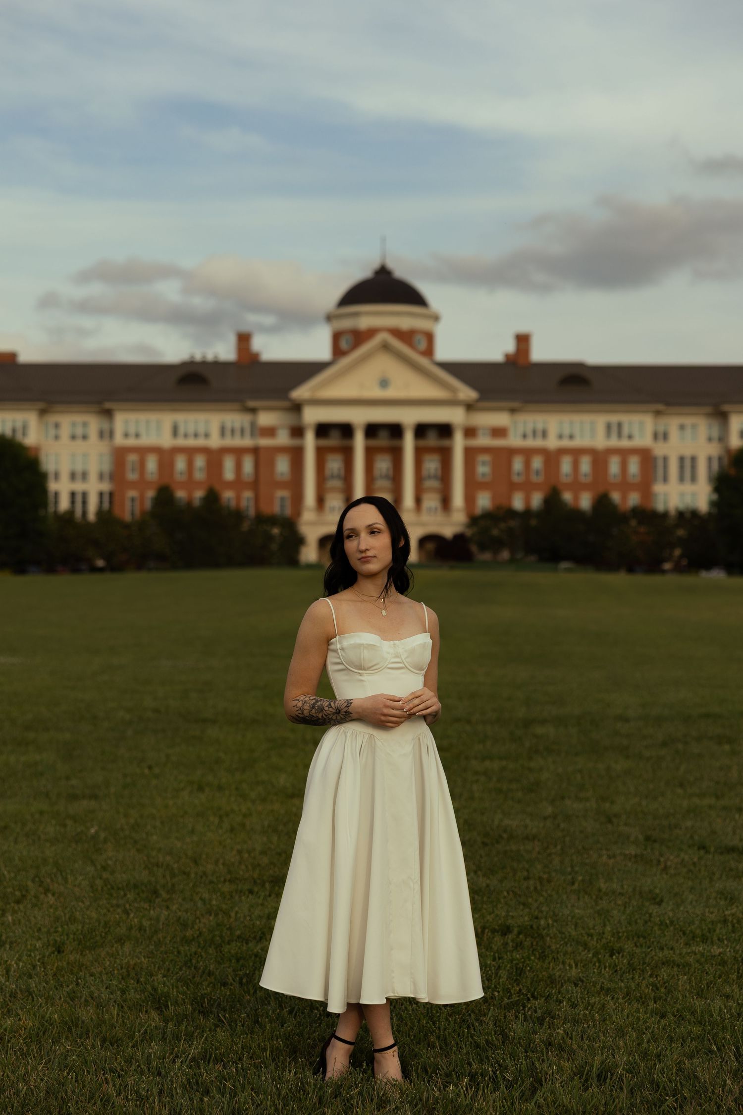 Woman in white midi dress with tattoos stands on green lawn before a grand brick colonial building at dusk.