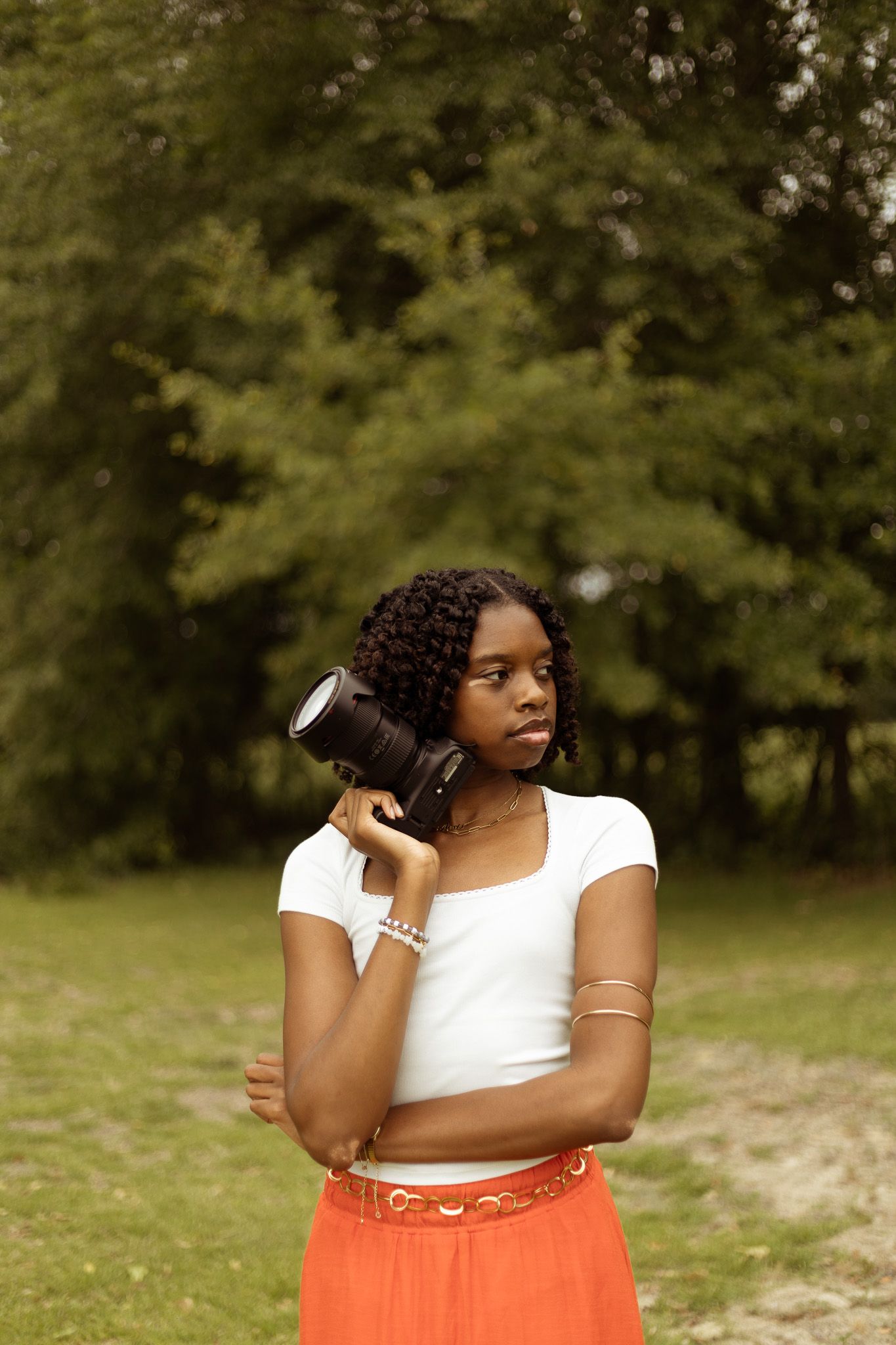 A person in a white top and orange skirt poses outdoors with headphones in a natural setting.