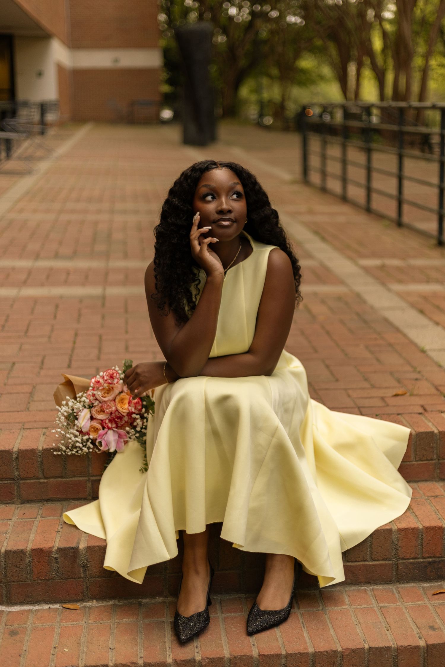 Young Black woman in yellow dress sits on brick steps holding colorful bouquet, posing thoughtfully outdoors.