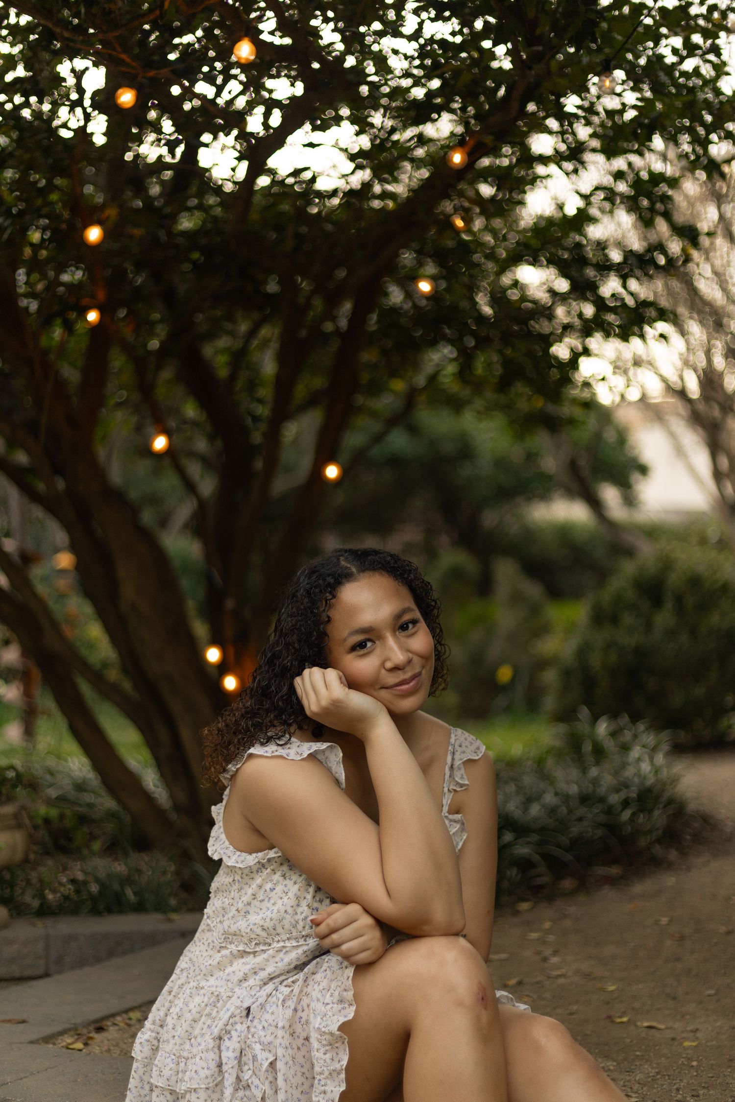 A person in a lace dress sits on the ground at sunset with string lights glowing in the trees above.