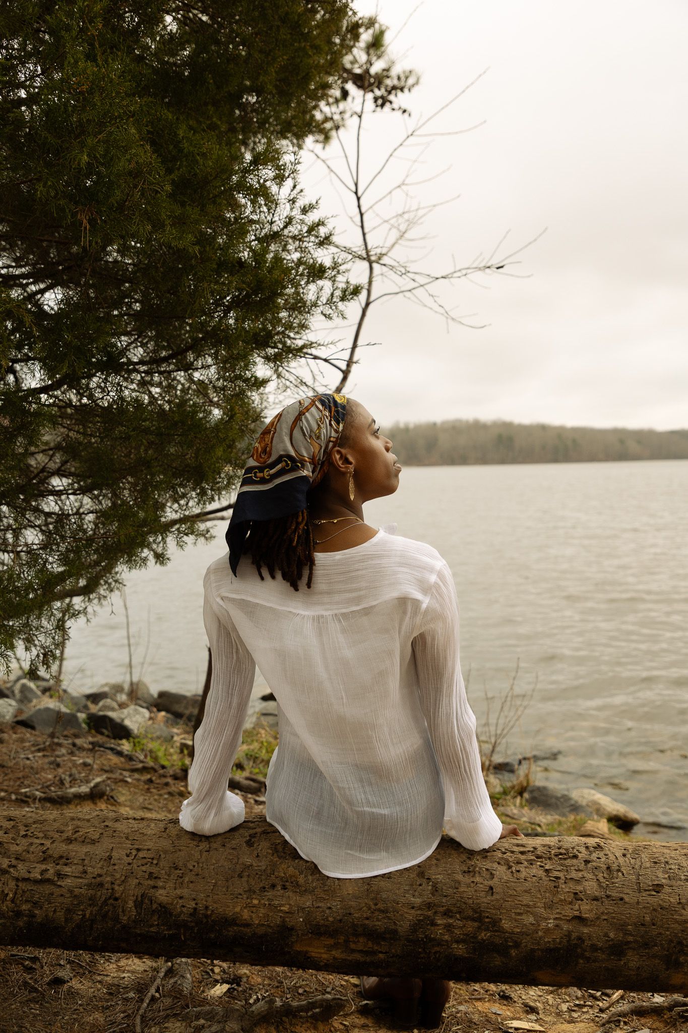 Person in white blouse sits on fallen tree trunk by peaceful lake at sunset, looking toward water.