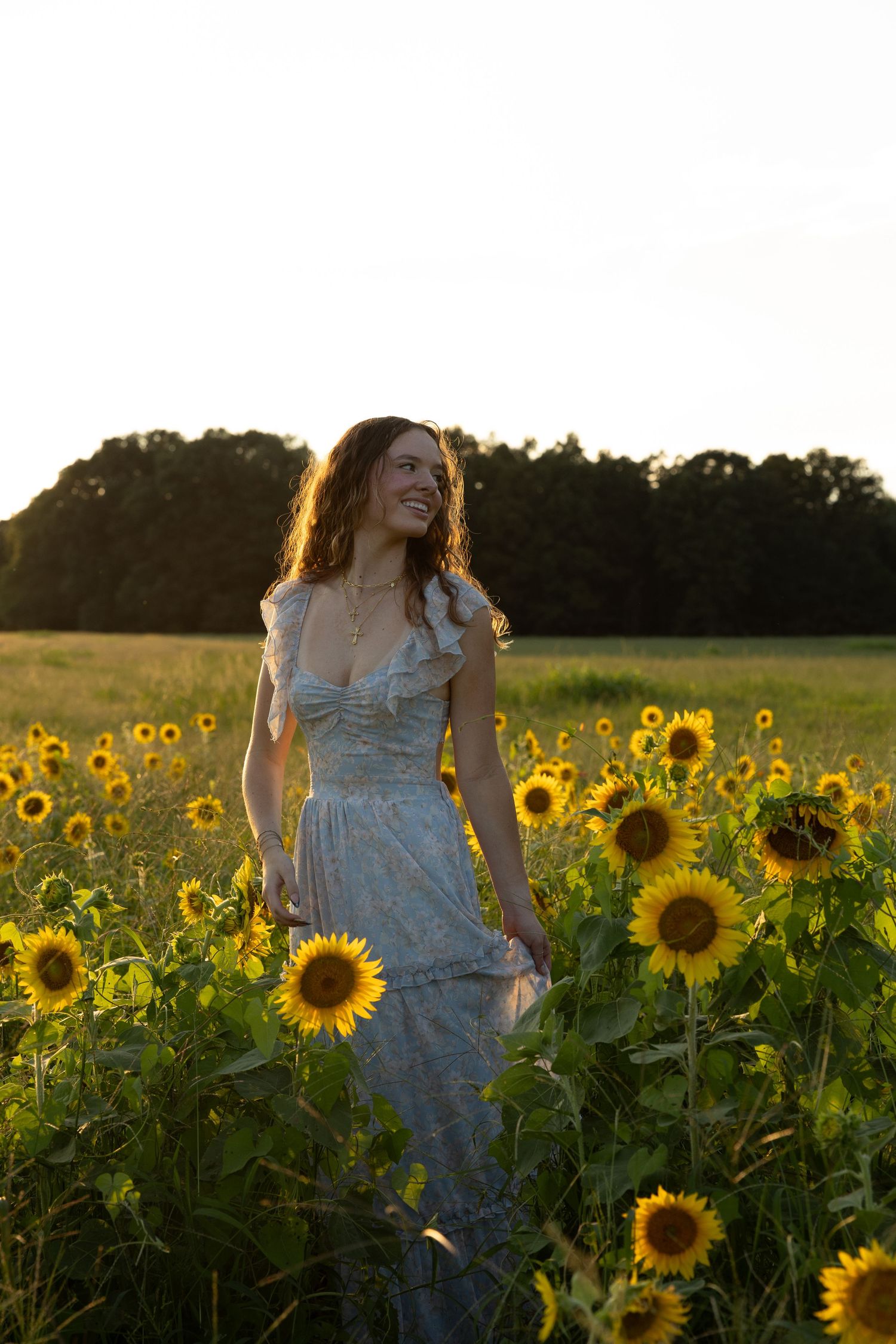 A figure stands amid vibrant sunflower fields at golden hour, wearing a flowy white summer dress against warm evening light.