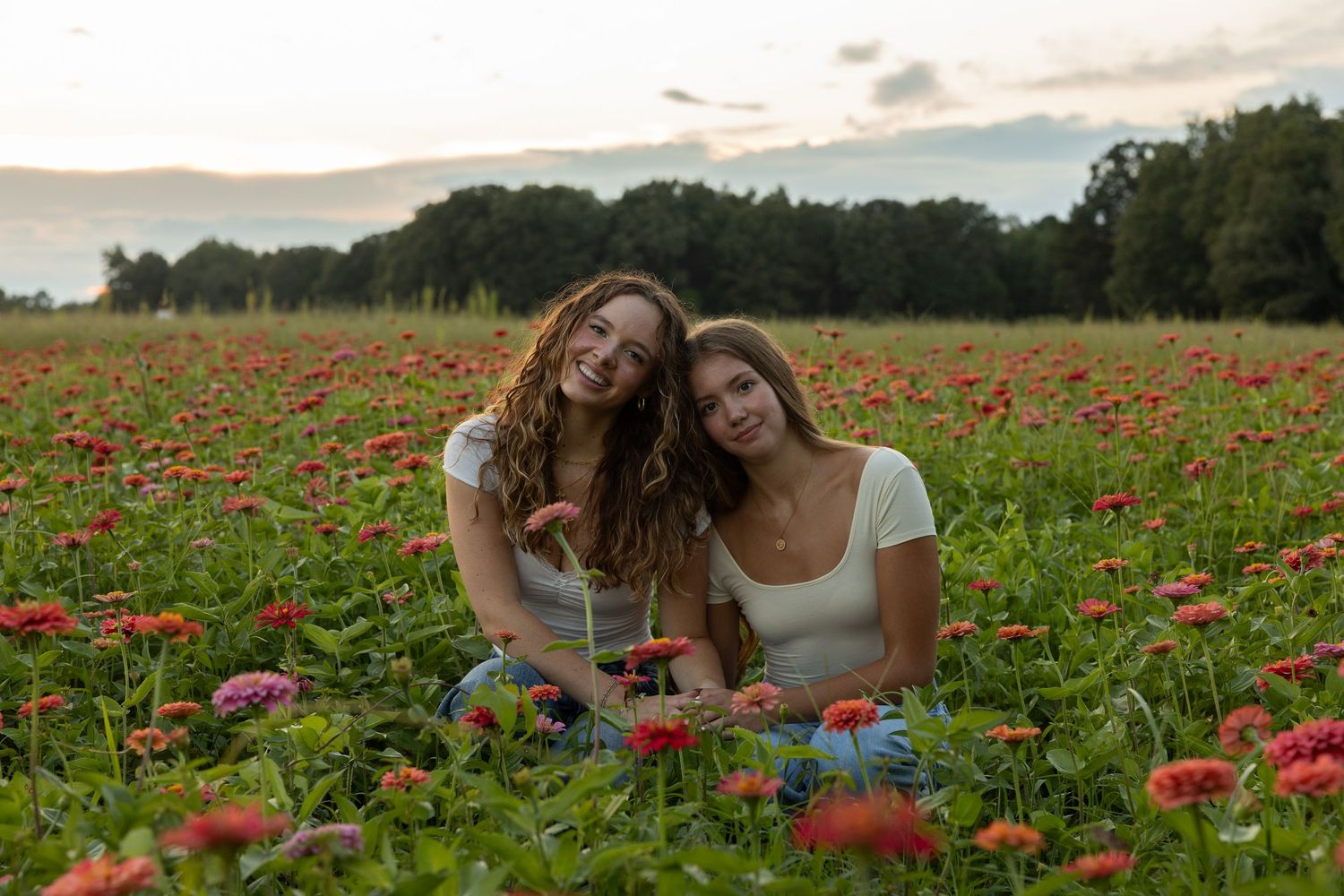 Two friends sitting together in a vibrant red poppy field at sunset with trees in the background.
