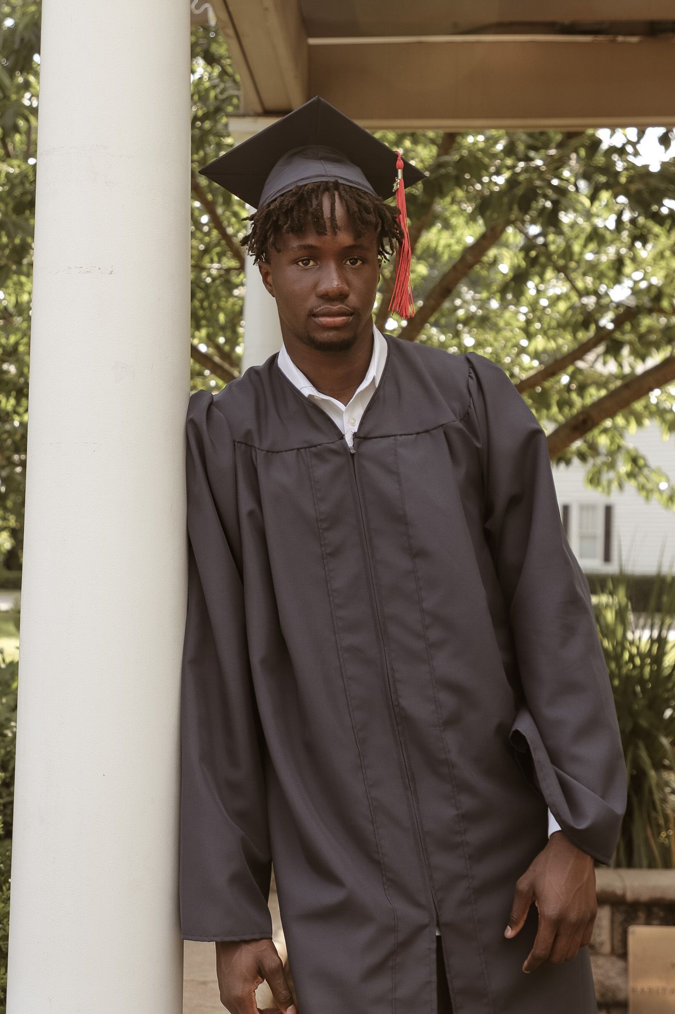 Person wearing graduation cap and gown poses near white columns in outdoor setting.