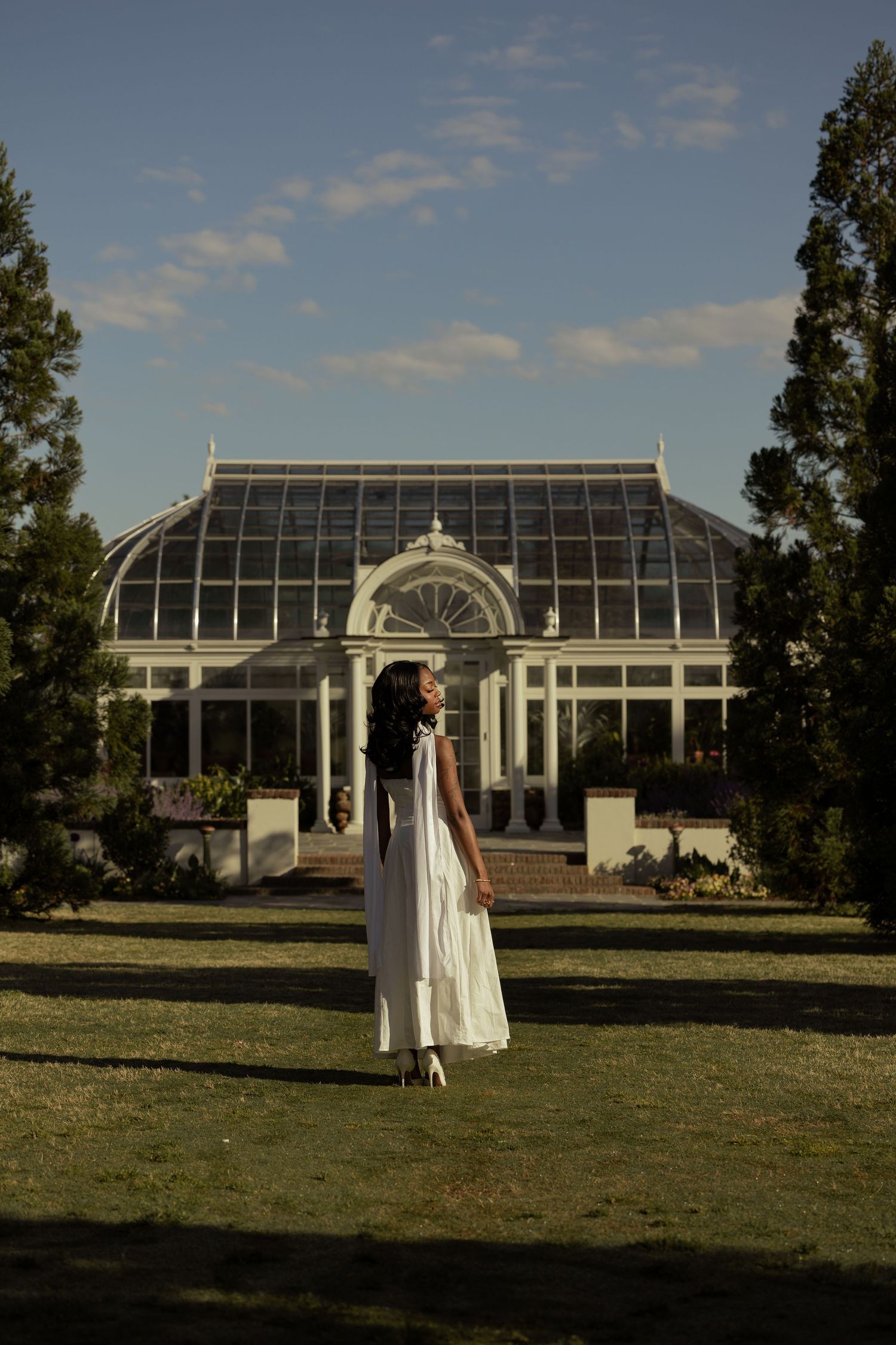 Woman in white maxi dress stands on green lawn before elegant Victorian greenhouse surrounded by tall trees.