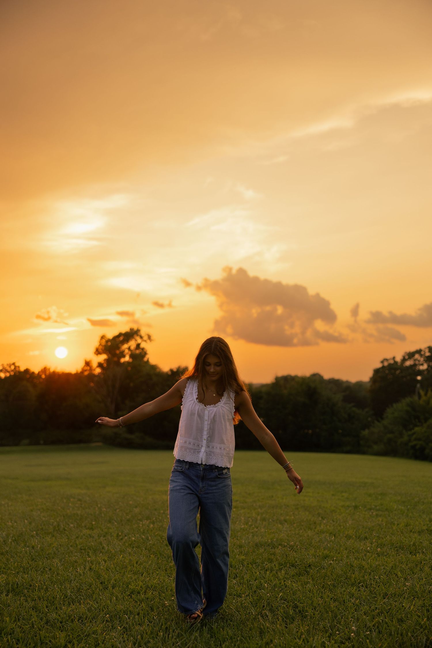 Woman in white top and jeans walks through green field at golden sunset with dramatic orange sky.