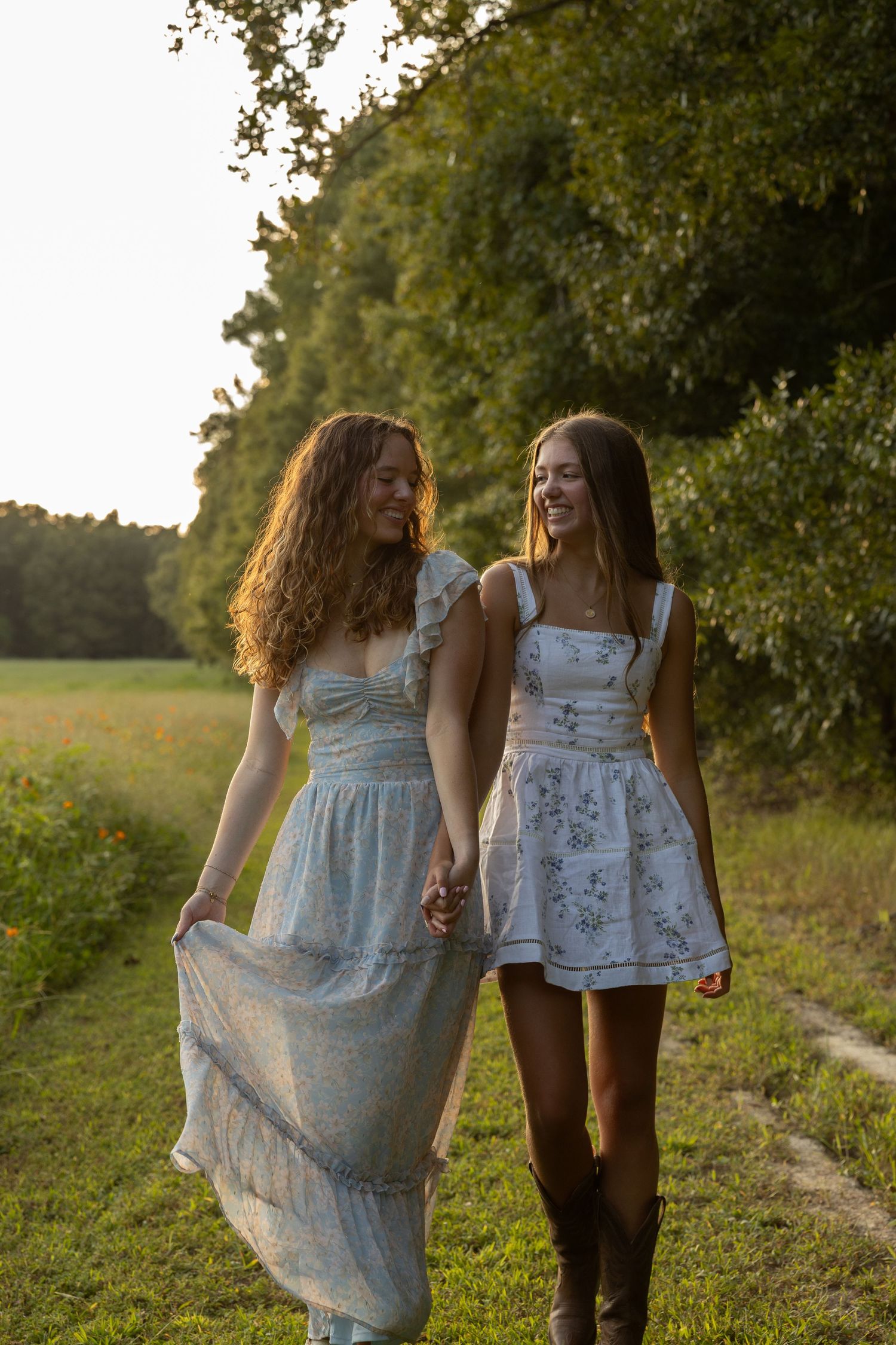 Two friends in summer dresses walk along a countryside path at golden hour, with sunlit trees in the background.