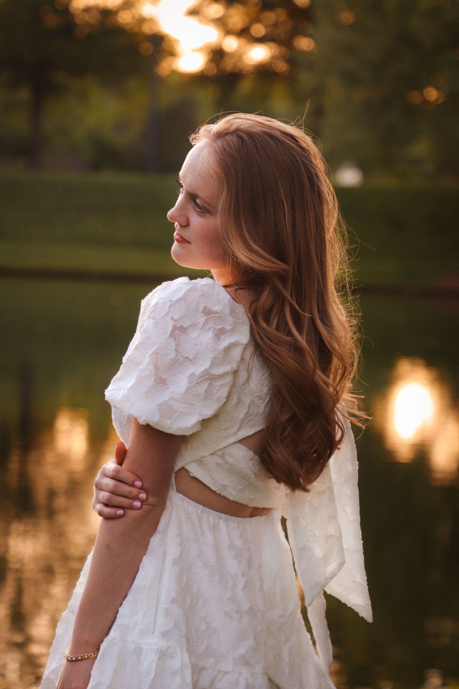 Woman in white dress poses at sunset by a reflective pond.