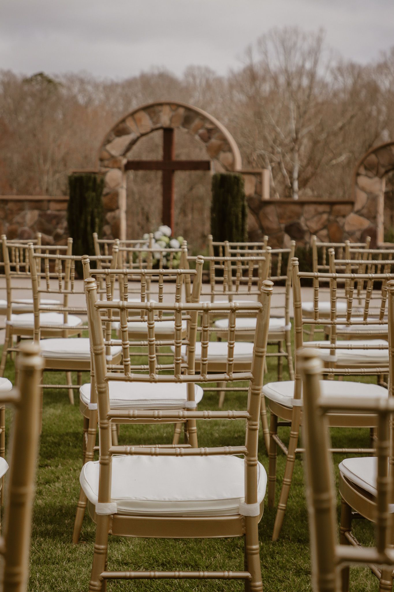 Golden chairs arranged for an outdoor wedding ceremony with a stone arch and cross backdrop.