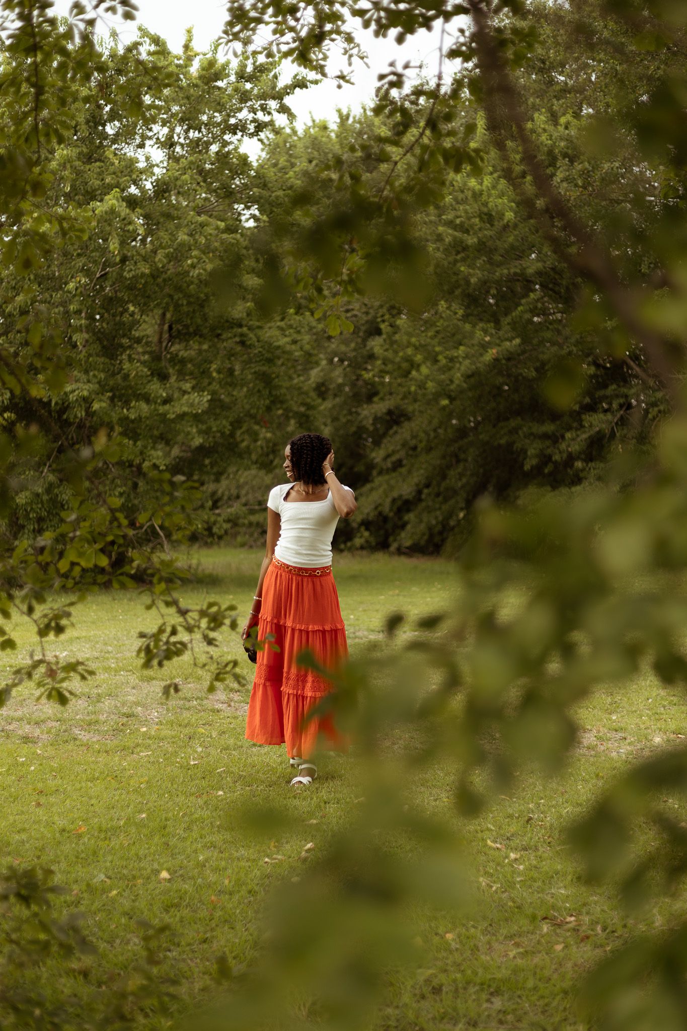 A person in a white top and flowing red skirt explores a lush green garden on a sunny summer day.