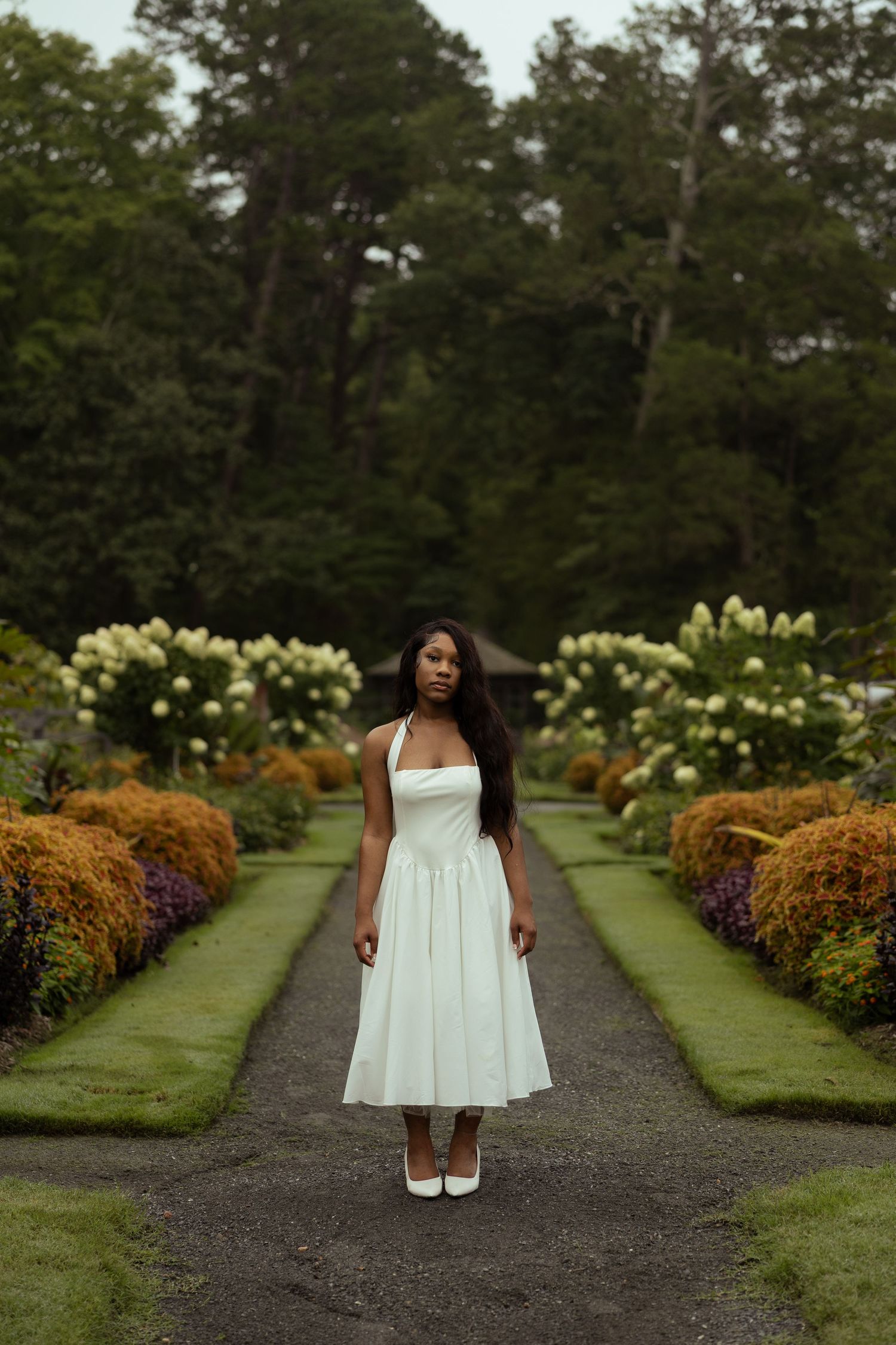 Woman in white halter dress and heels stands on garden path surrounded by colorful flowers and lush green trees.