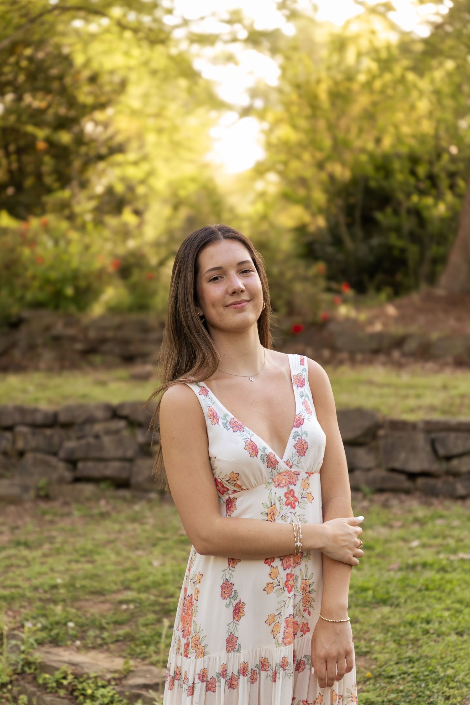 Person in floral sundress poses outdoors at sunset in a garden setting with stone wall and greenery.