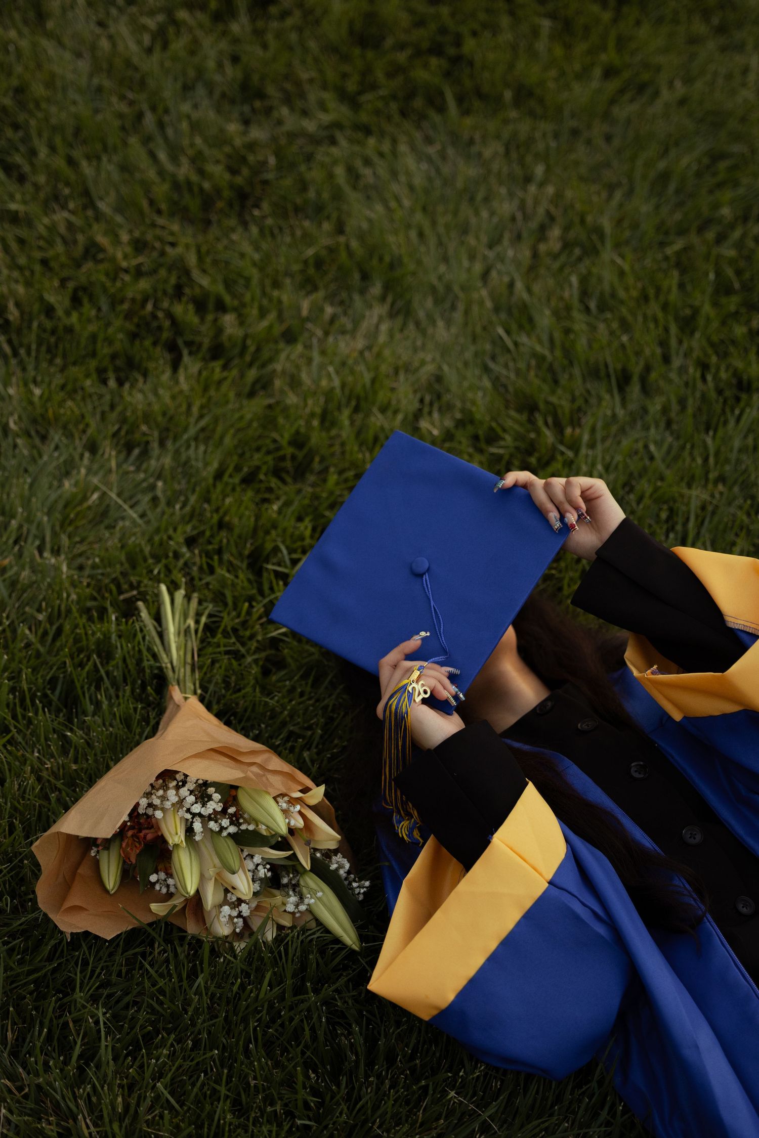 Graduate in blue and gold cap and gown lying on grass holding mortarboard beside a wrapped flower bouquet.