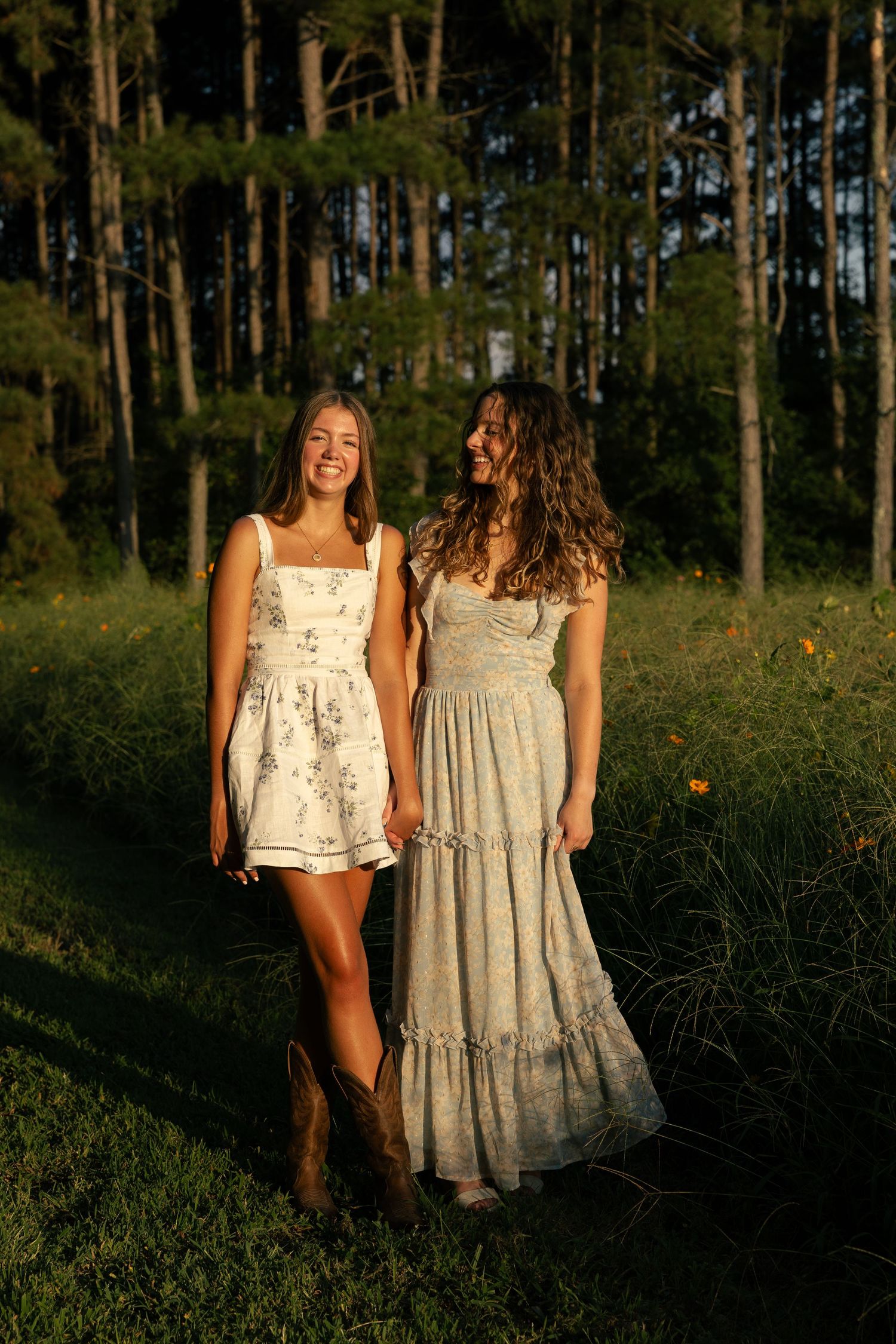 Two young women in floral dresses and cowboy boots smile together in a sunlit meadow with tall pine trees behind them.