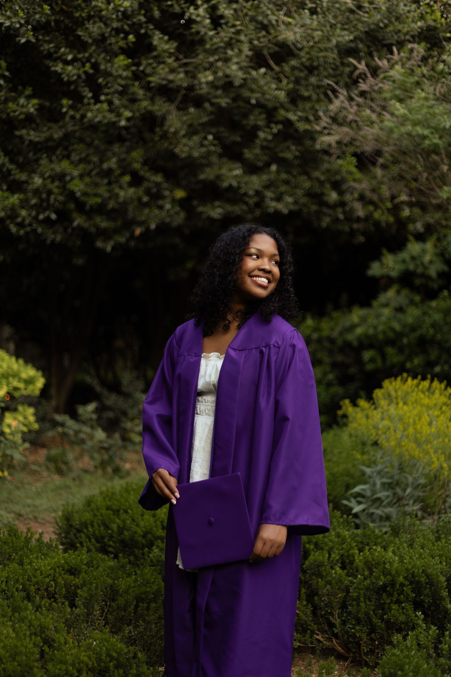 Smiling young woman in purple graduation cap and gown standing outdoors among green shrubs and trees.