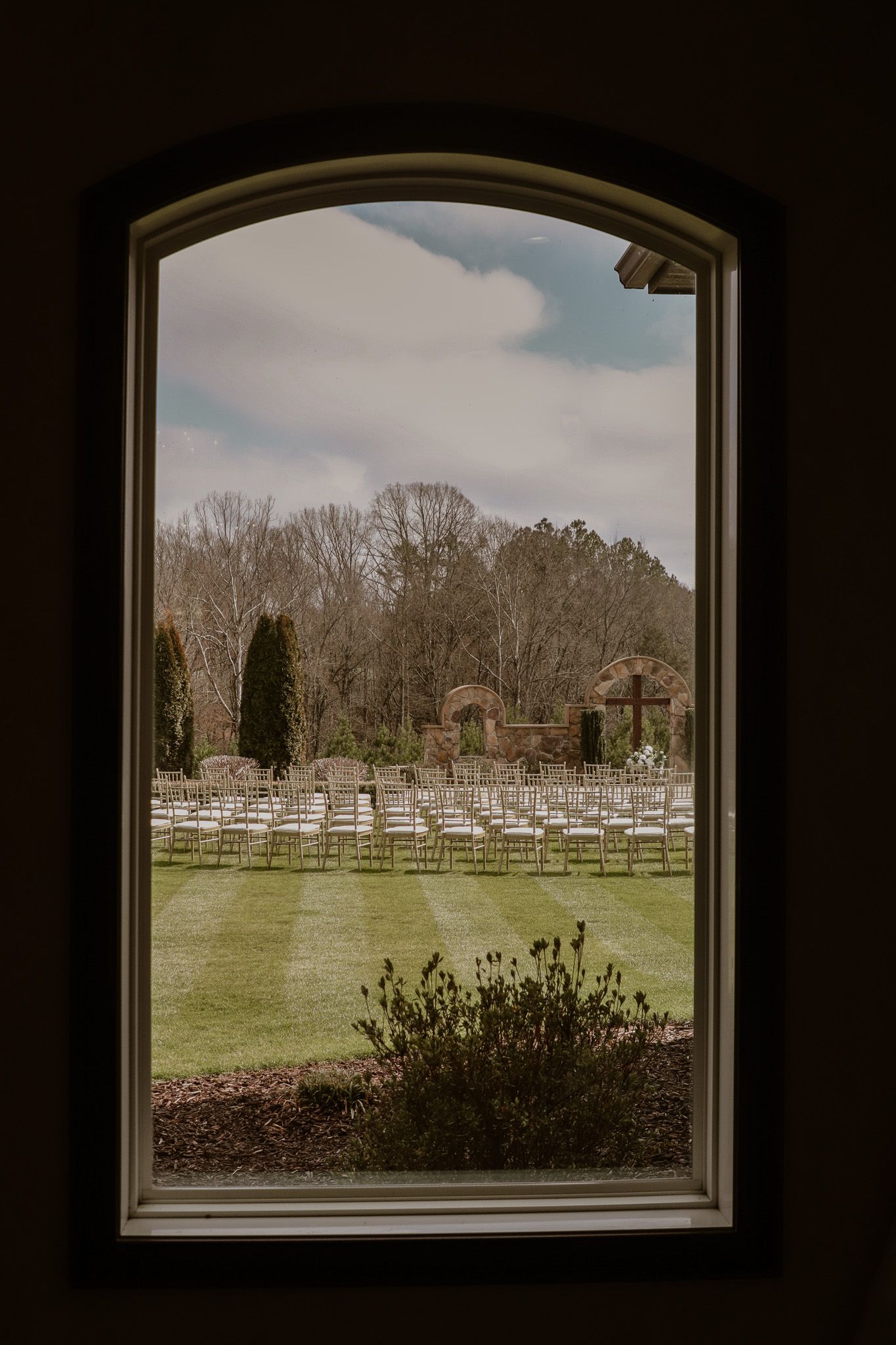 Wedding ceremony setup with white chairs viewed through an arched window.