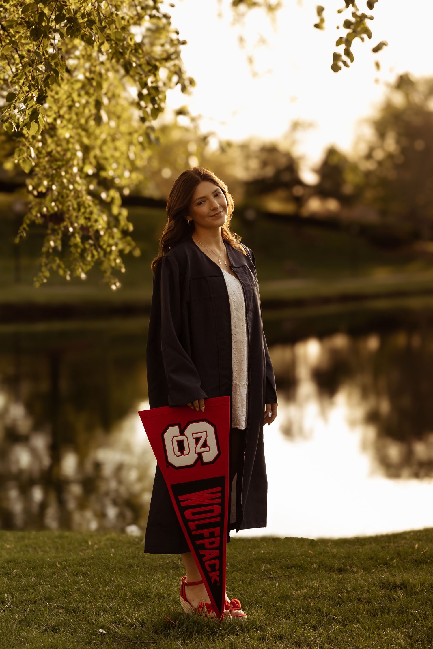 A person in a black coat holds a red banner while posing by a serene lake at sunset, creating a tranquil outdoor scene.