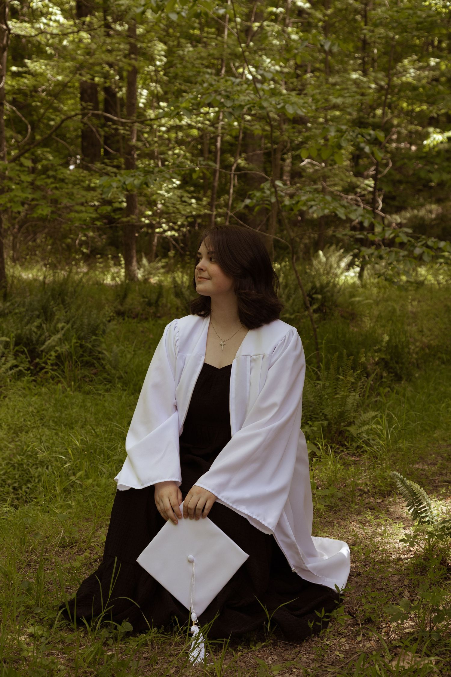 A person in a flowing white cardigan and black dress sits on grass in a peaceful forest setting.