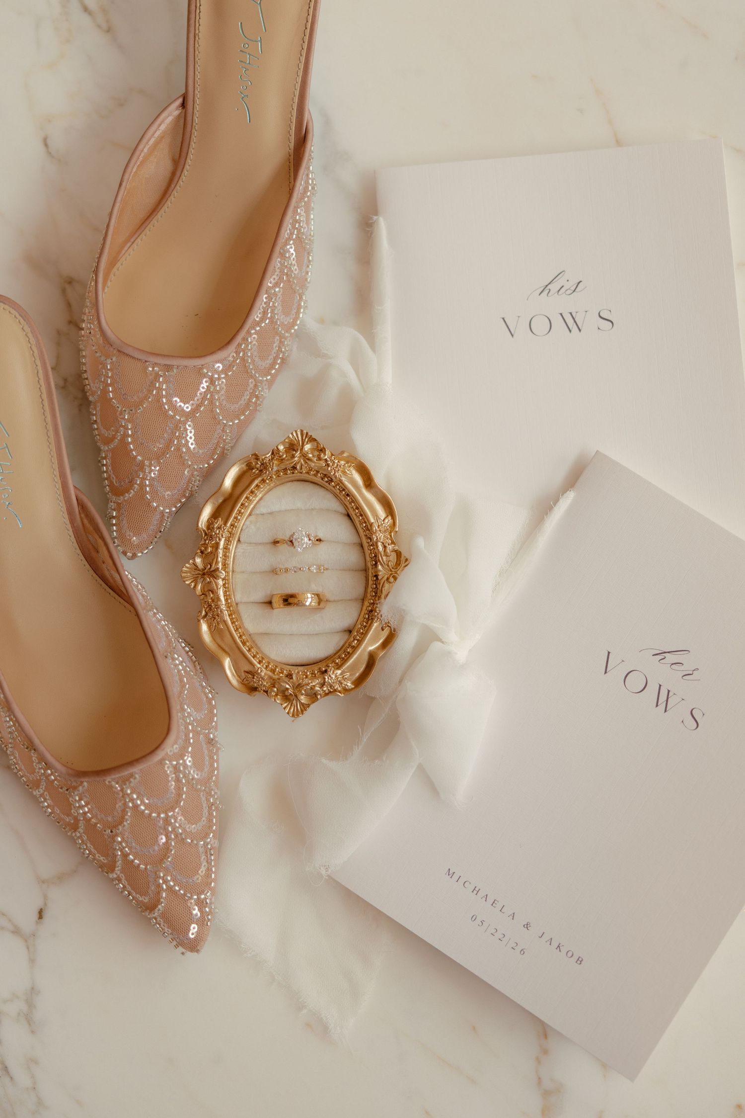 Elegant wedding flat lay with beaded blush heels, gold rings in ornate frame, and his and her vows booklets on marble.