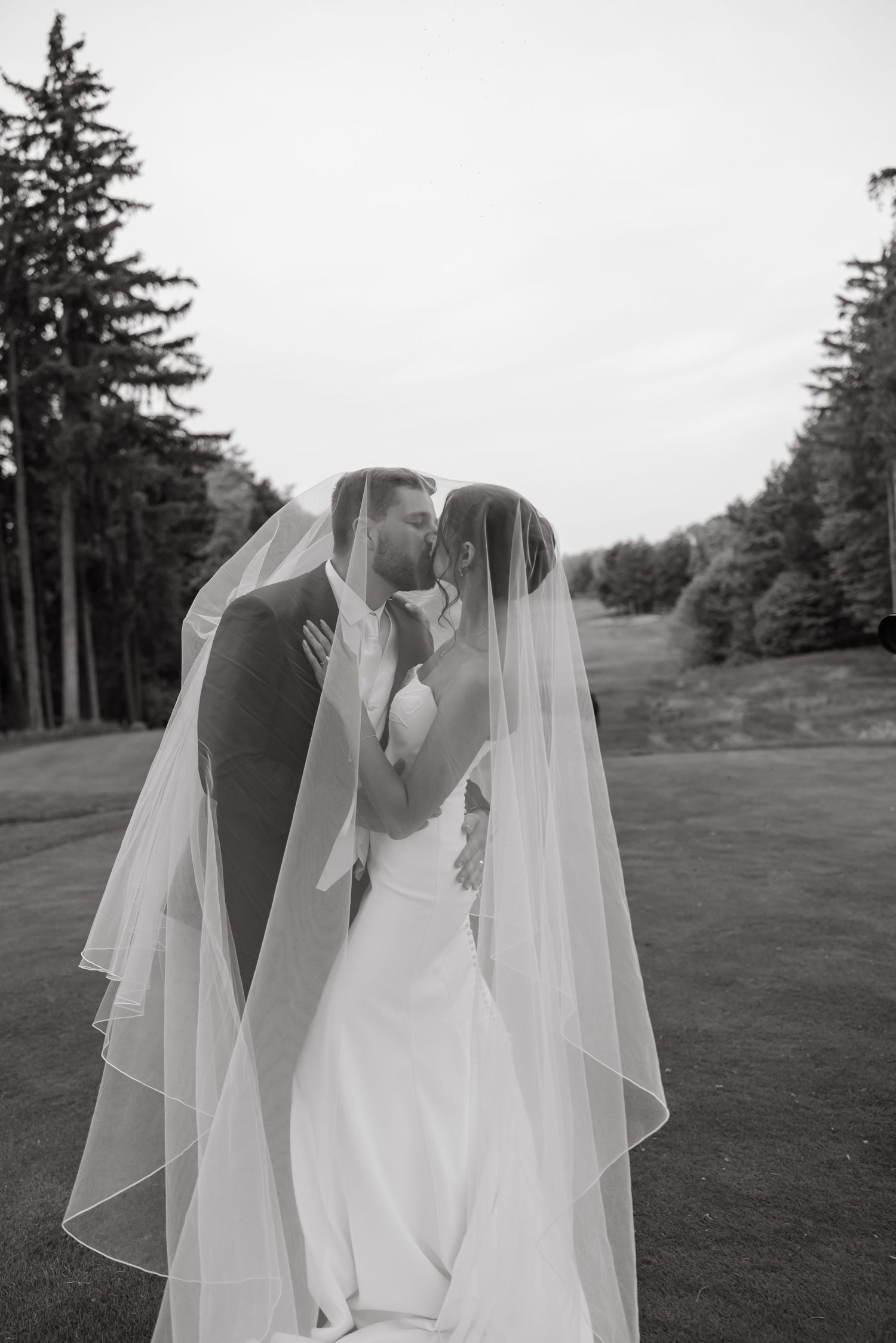 Black and white photo of a bride and groom kissing under a flowing veil outdoors, surrounded by tall evergreen trees.