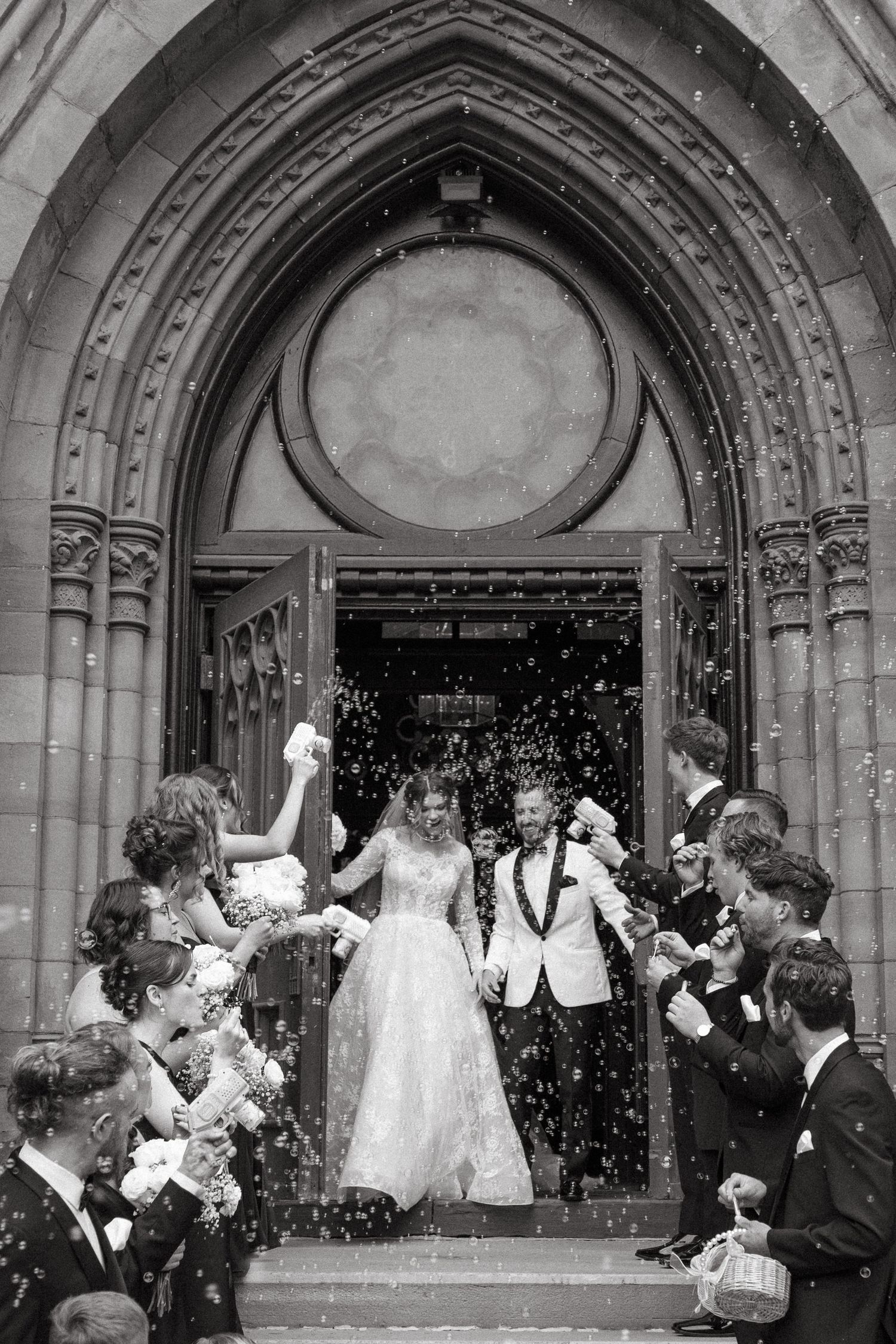 Wedding party exits church through stone archway as guests throw confetti and celebrate the joyous occasion.