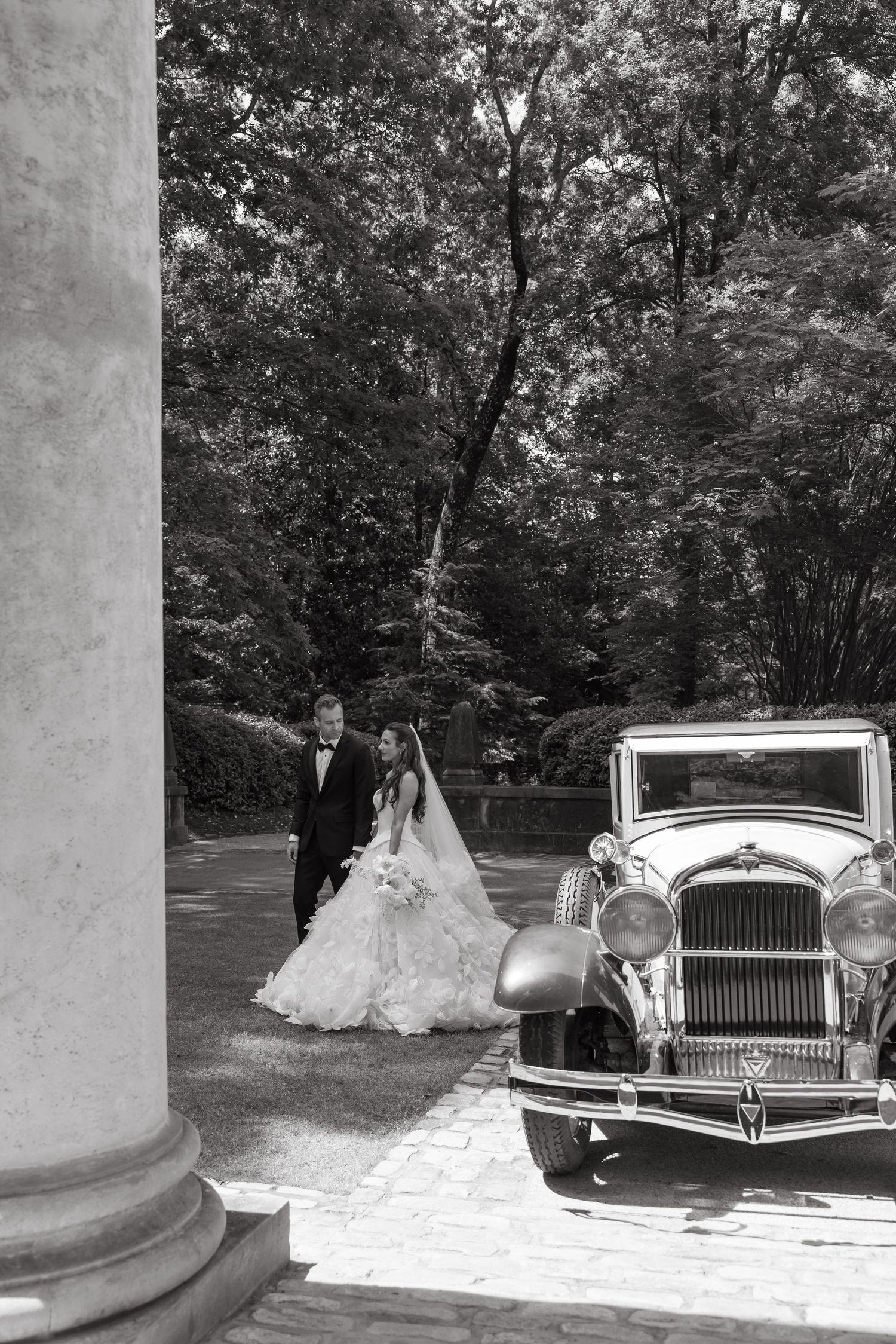 Black and white photo of bride and groom walking toward a vintage classic car amid lush trees and elegant columns.