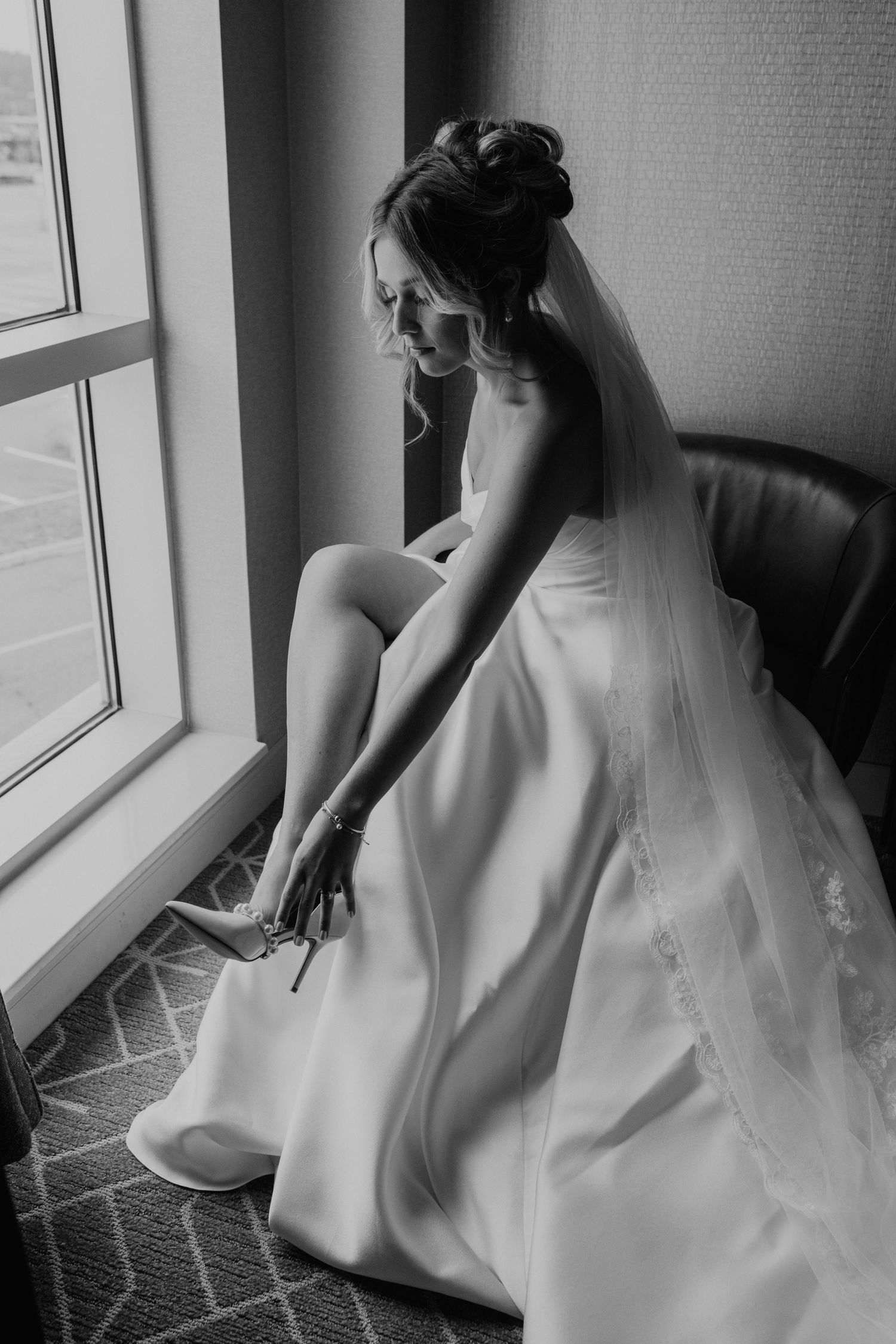 Black and white photo of bride adjusting her shoe while seated by window in flowing wedding dress.