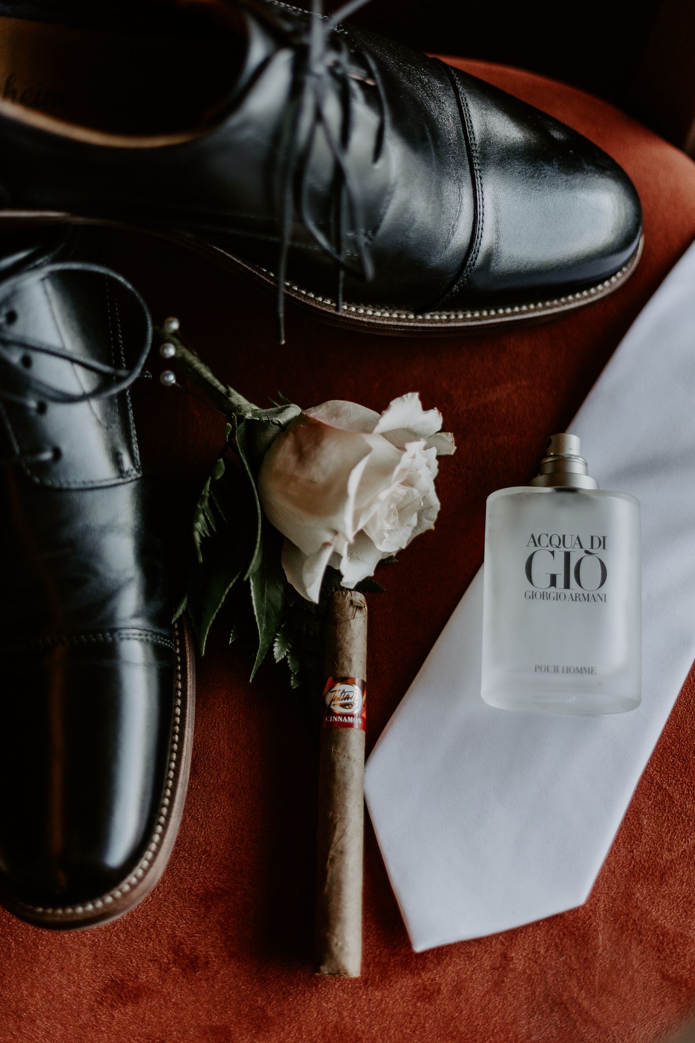 Black leather dress shoes with a white rose boutonniere and Acqua Di Gio cologne arranged on a rust-colored surface.
