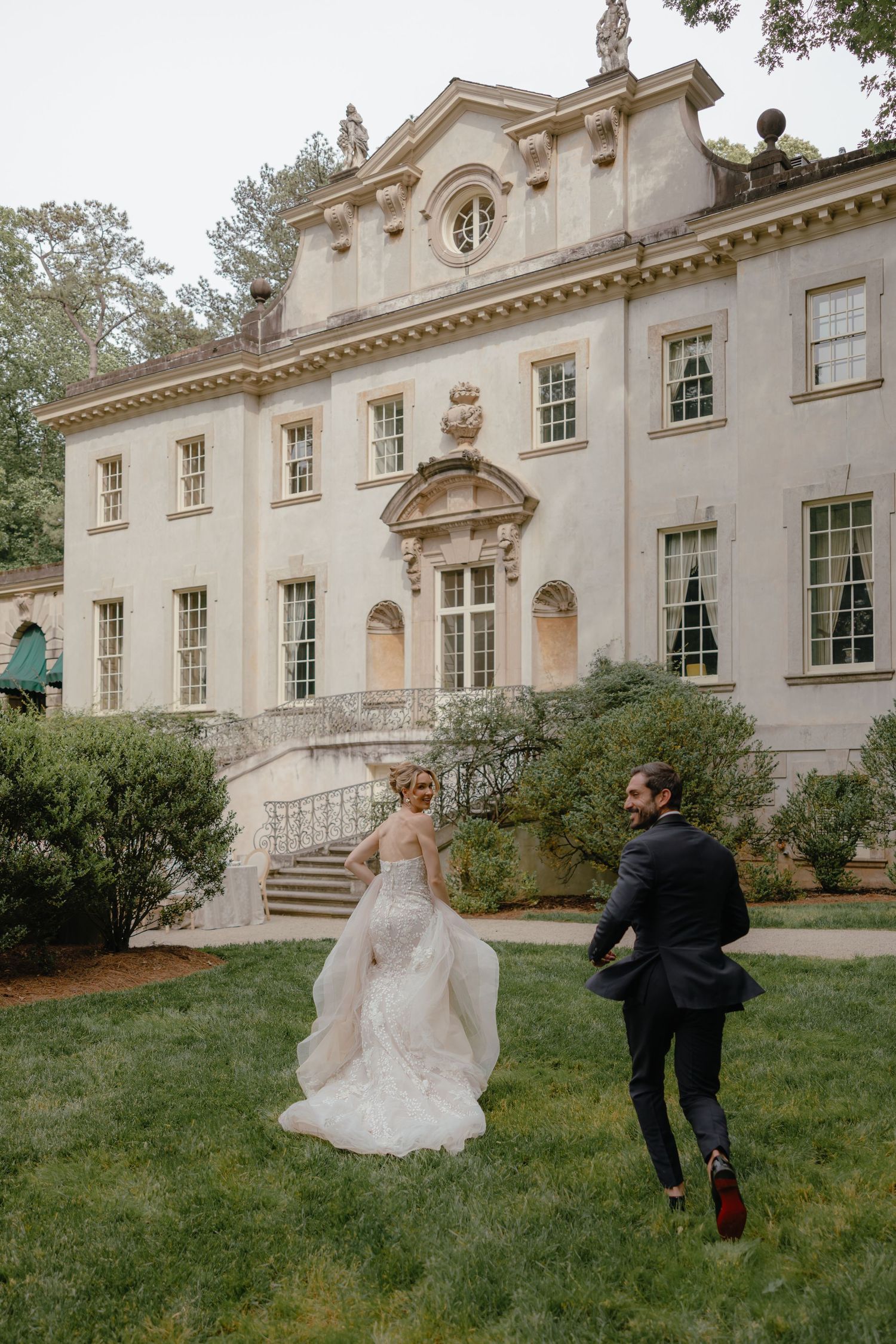 Bride and groom running across lush green lawn in front of an elegant historic mansion wedding venue.