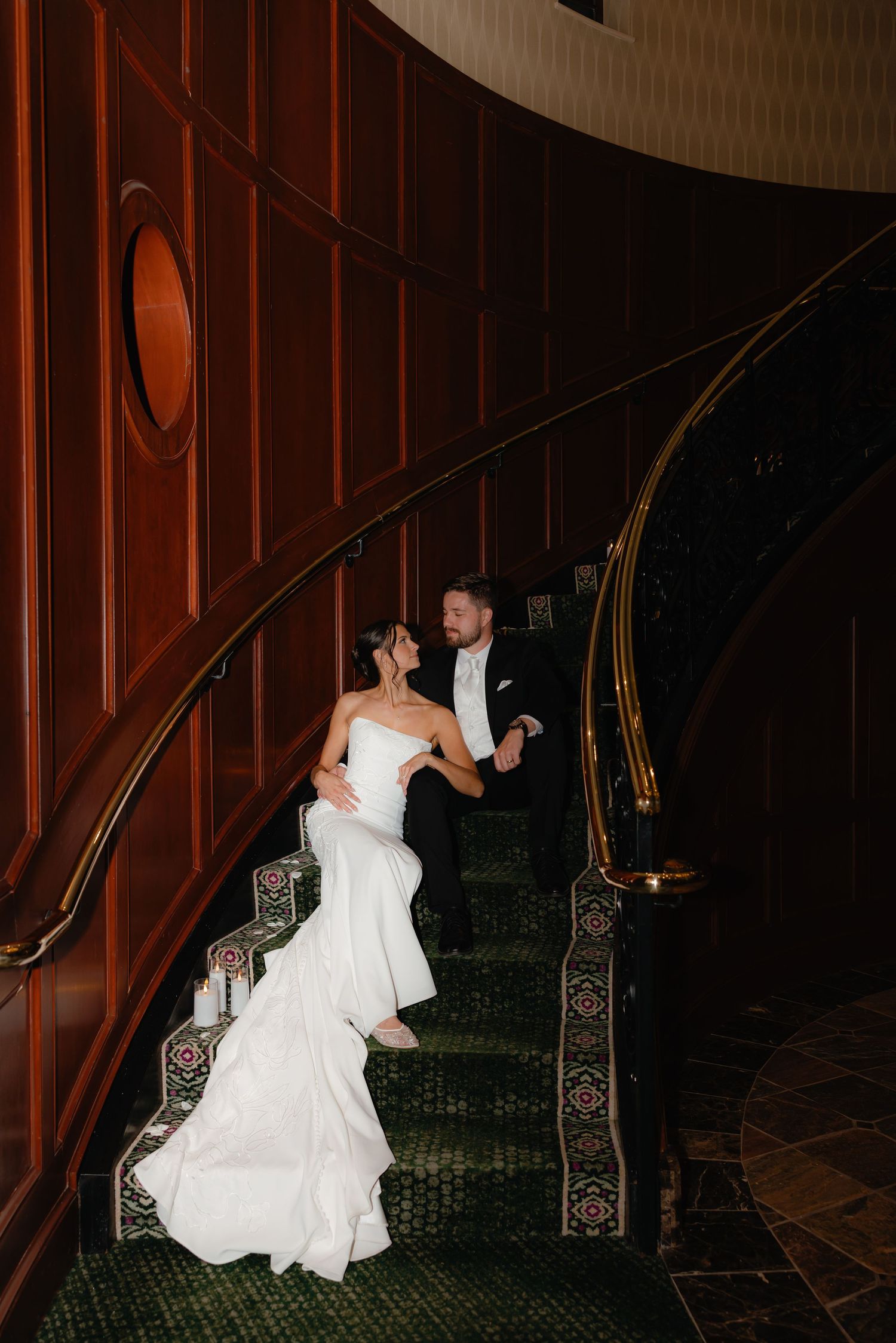 Bride and groom share a romantic moment on an elegant curved staircase with dark wood paneling and green carpet.