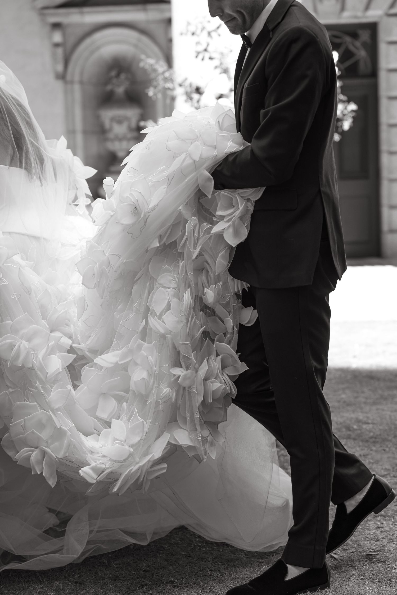 Black and white photo of groom in suit carrying bride's voluminous ruffled wedding dress train outdoors.