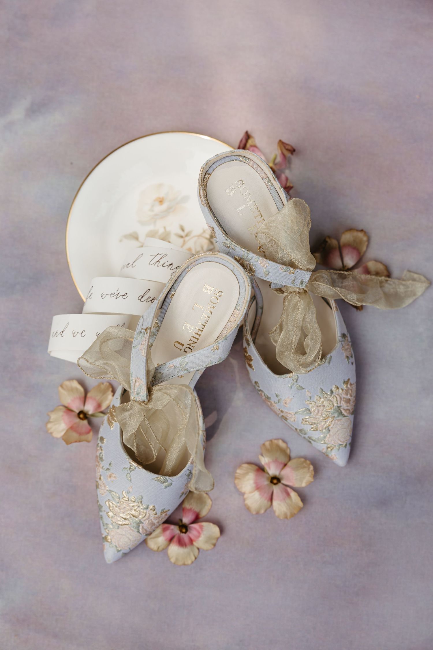 Elegant embroidered bridal heels in soft blue with gold lace details, organza bows, and pink flowers on a lavender background.