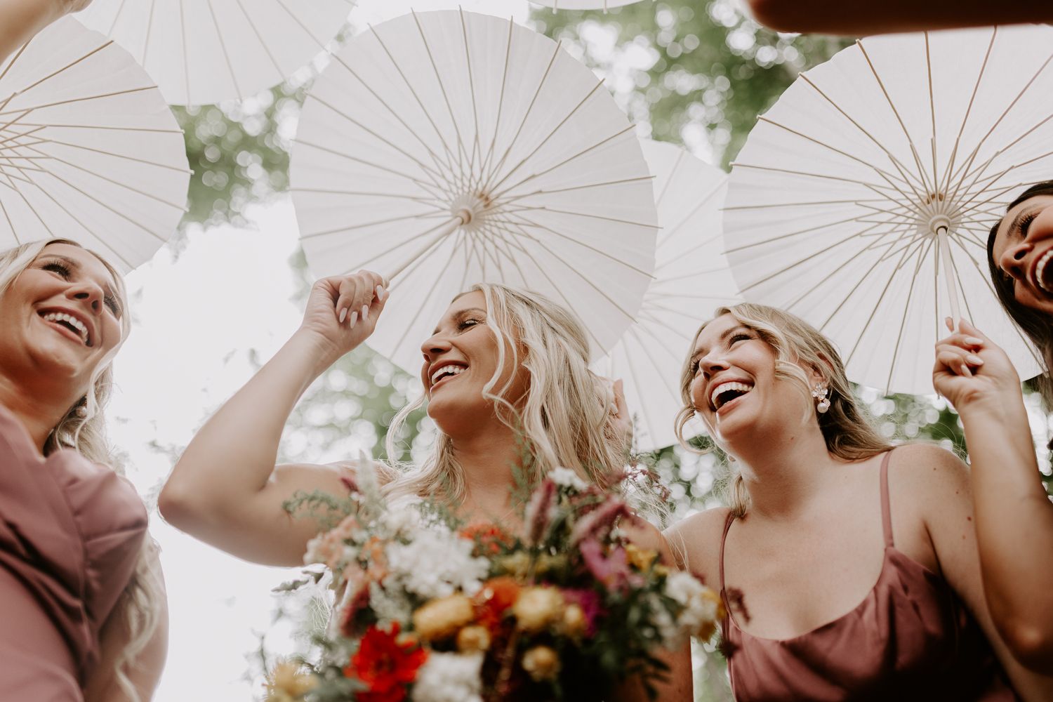 Wedding guests holding white parasols laugh joyfully during an outdoor celebration.
