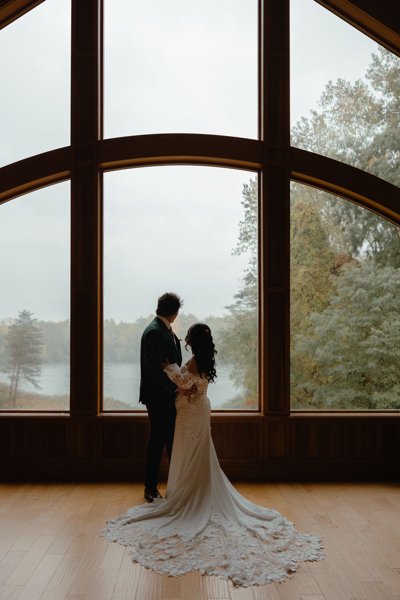 A wedding couple shares a tender moment silhouetted against large windows overlooking a misty forest landscape.
