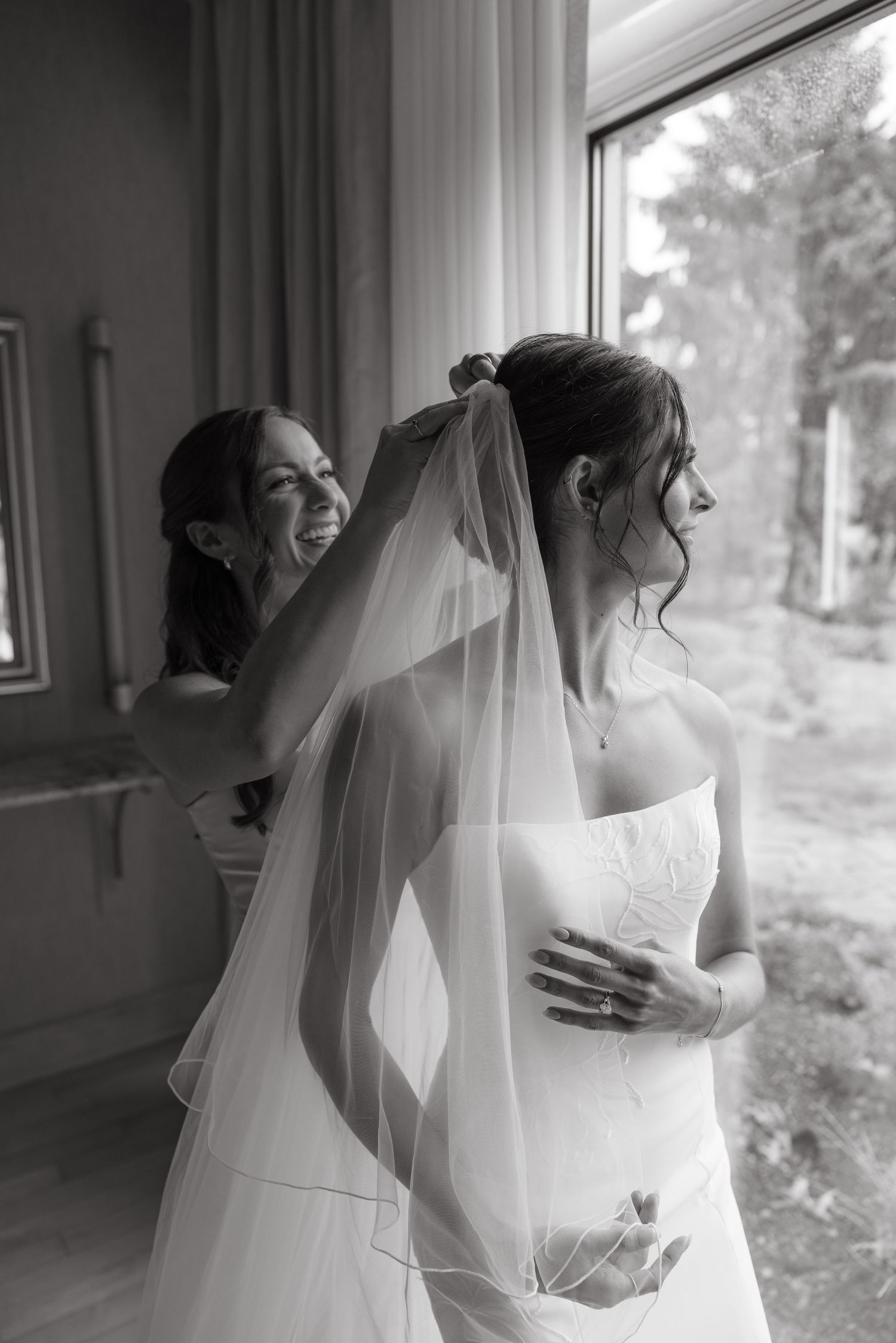Black and white photo of a smiling bridesmaid helping a bride put on her veil near a bright window.