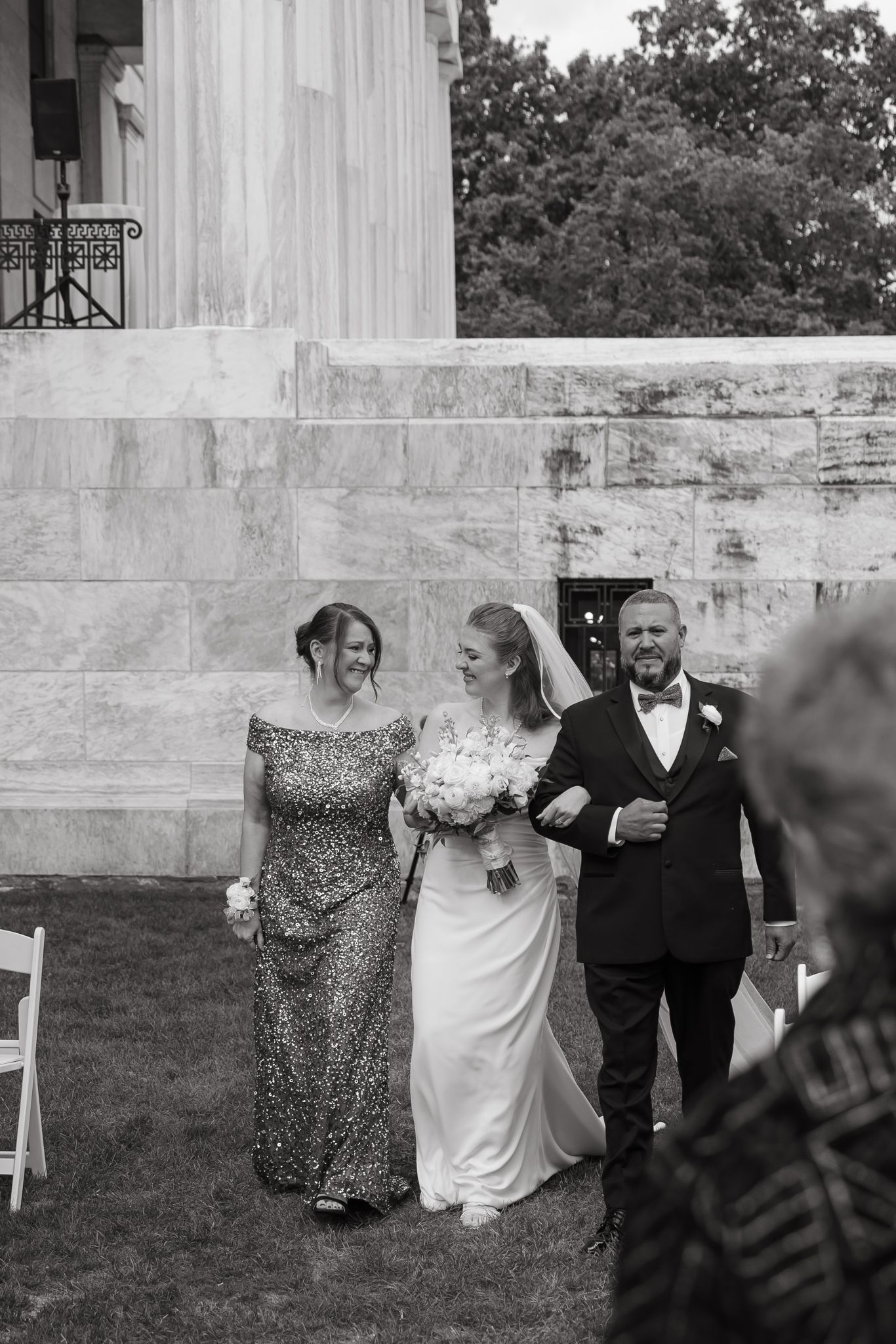 Wedding party members walk in a line outside a white stone building during an outdoor ceremony.