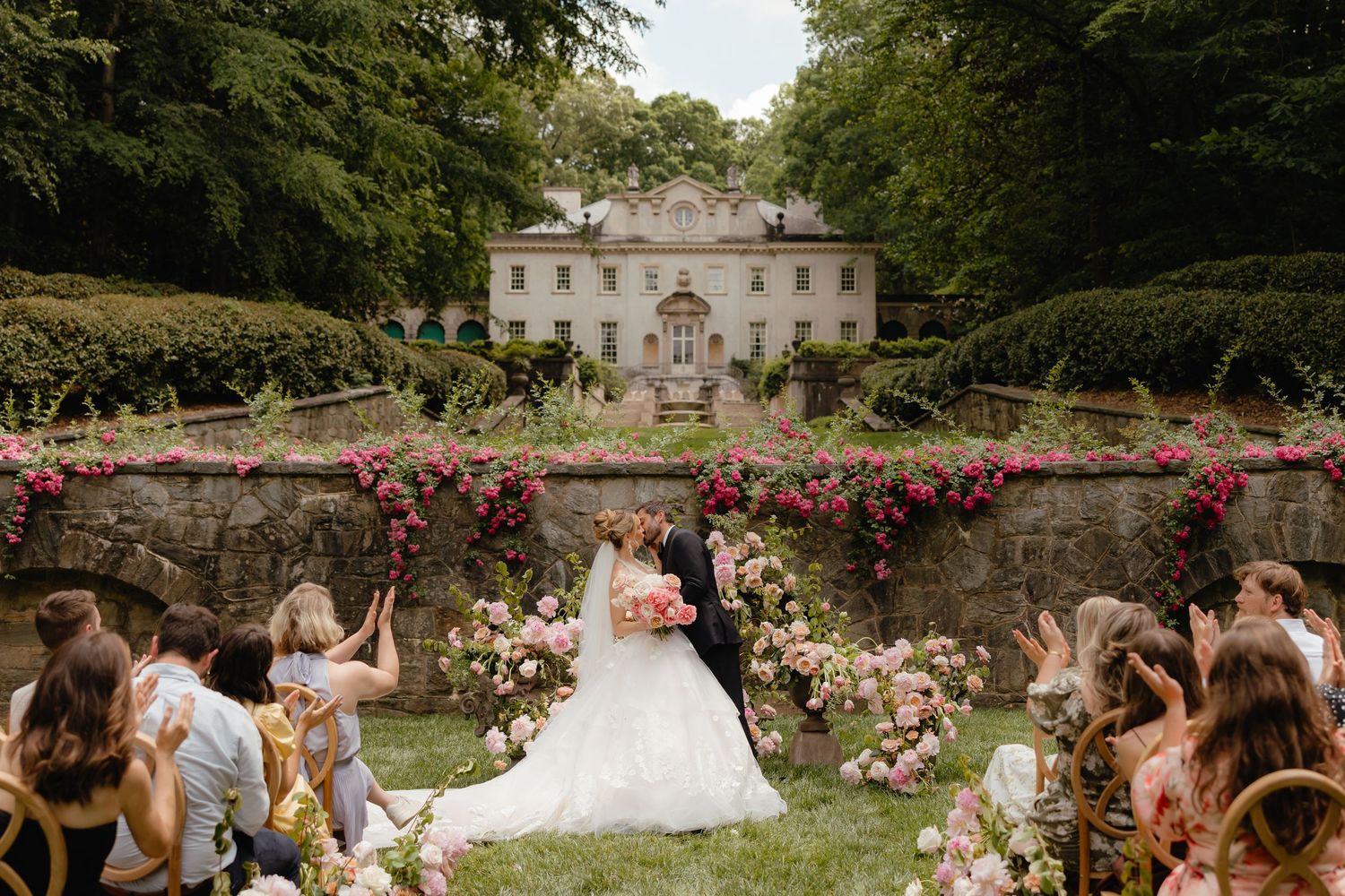Bride and groom kiss at outdoor wedding ceremony with floral arch, guests seated, and elegant manor house backdrop.