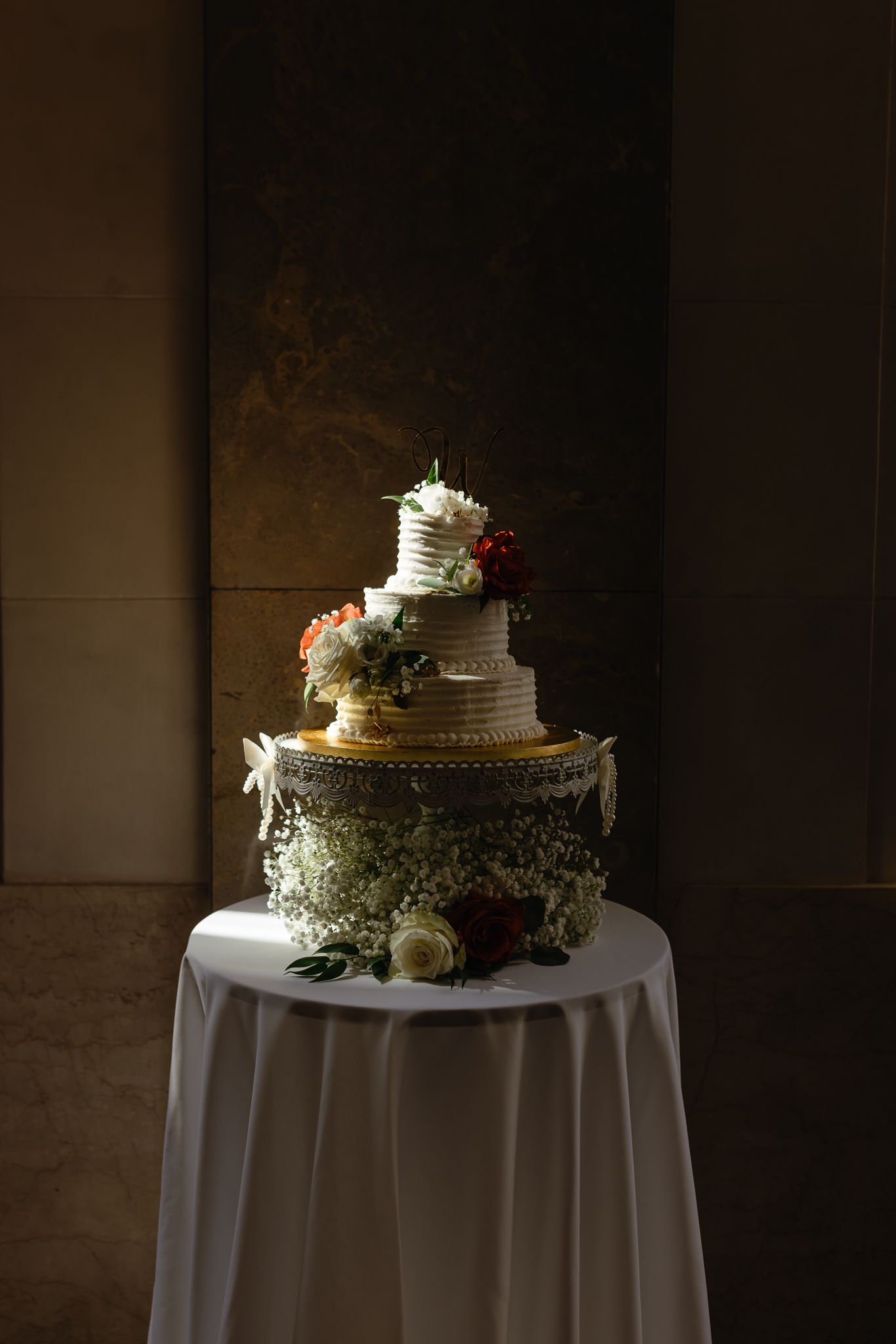 A white wedding cake with rustic decorations sits atop a round table with white tablecloth in dramatic lighting.