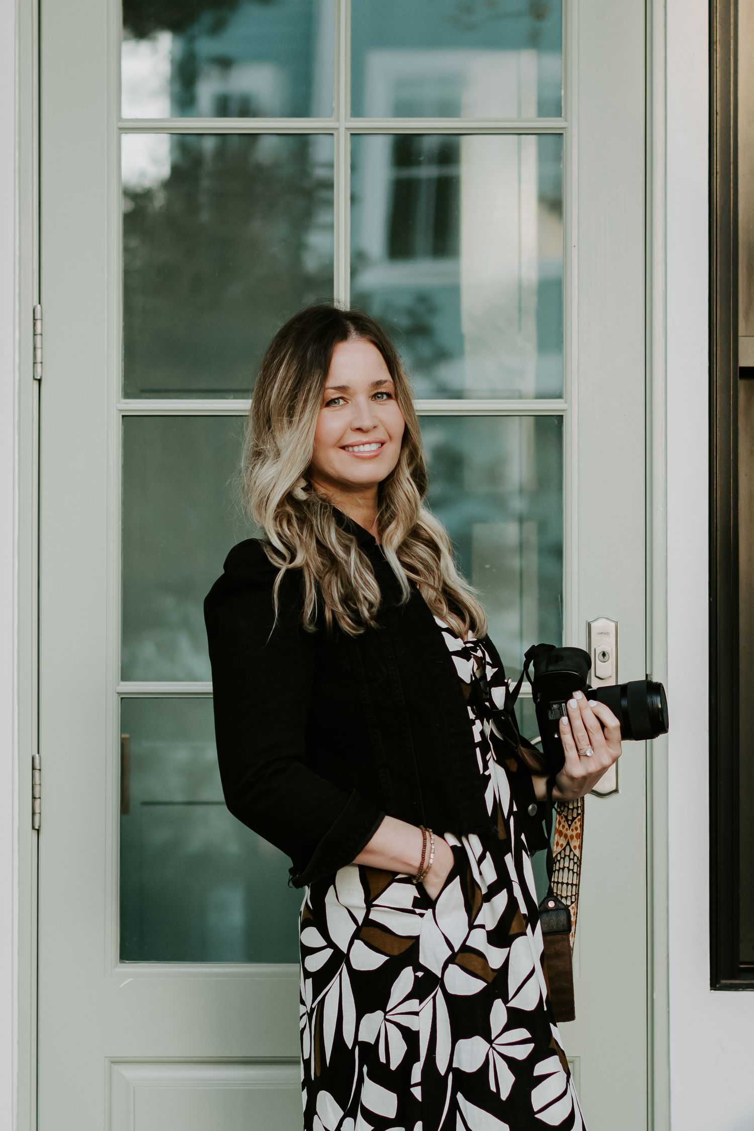 Professional photographer in floral dress holding camera against white door backdrop.