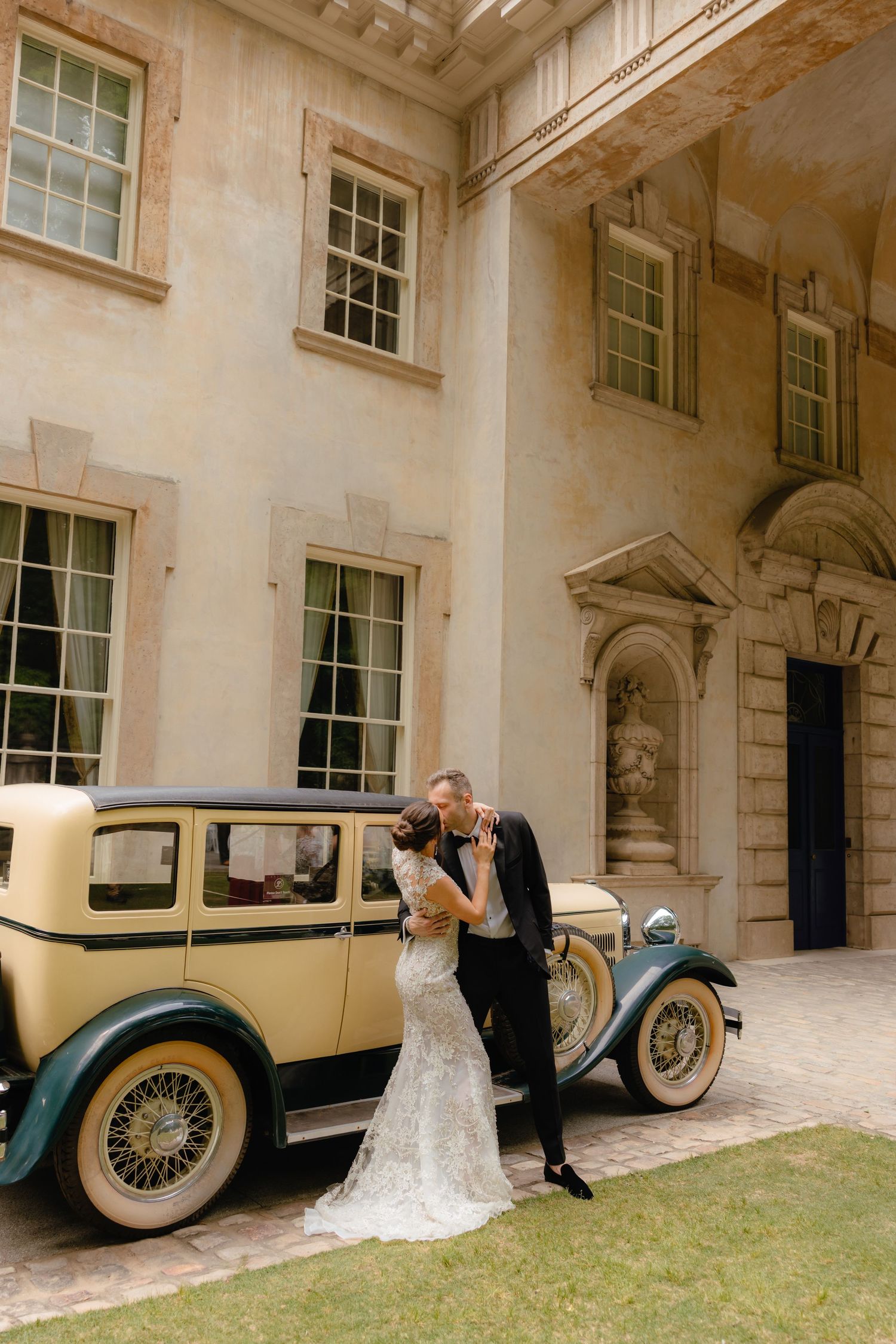 Bride and groom share a romantic kiss beside a vintage classic car outside an elegant historic mansion.