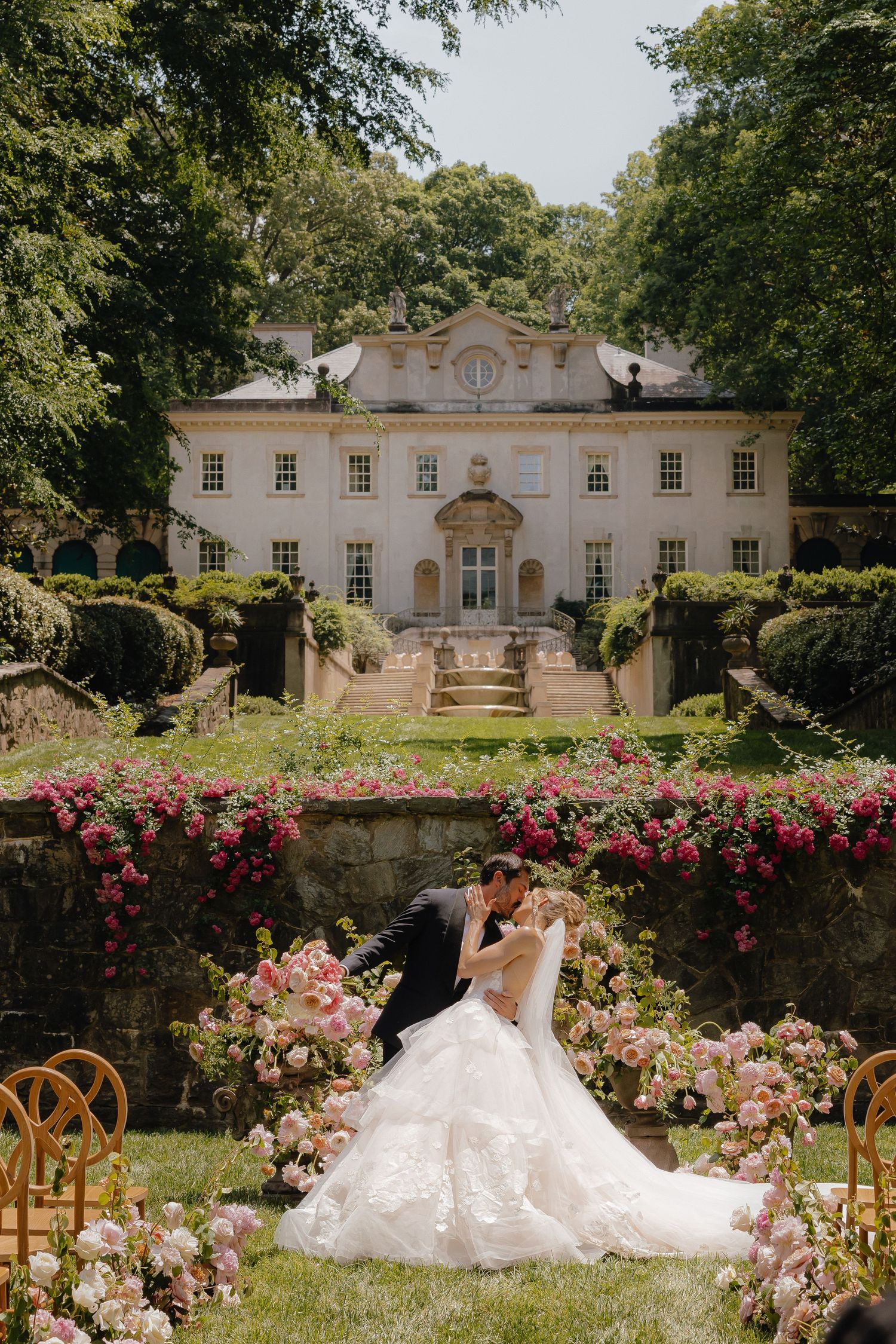 Couple kissing at outdoor wedding ceremony with floral arch, lush gardens, and elegant white mansion backdrop.