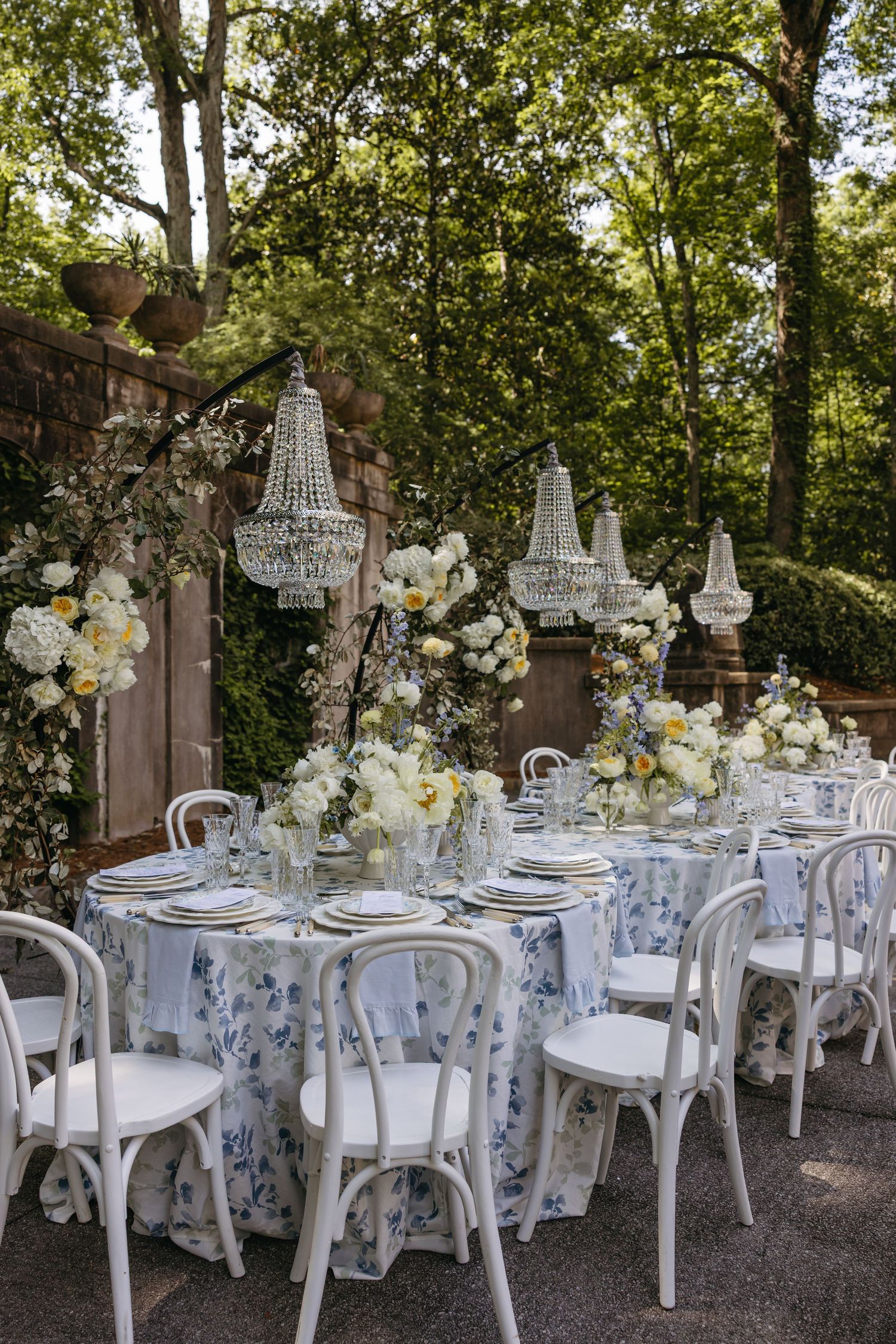 Elegant outdoor wedding reception with blue floral tablecloths, crystal chandeliers, white chairs, and lush floral centerpieces.