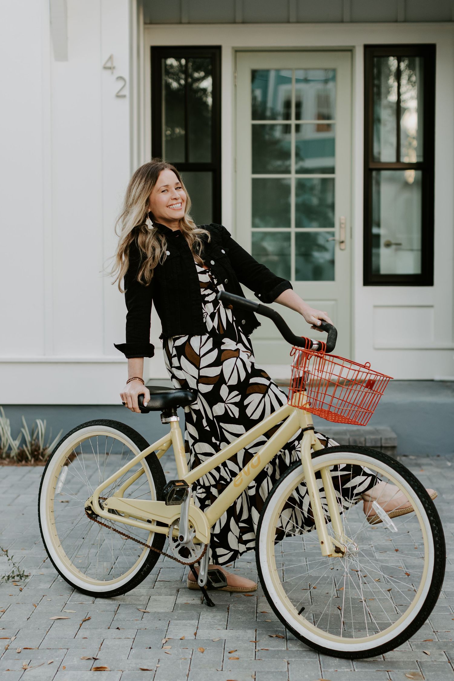 Woman in floral dress posing with cream-colored vintage bicycle outside white building.