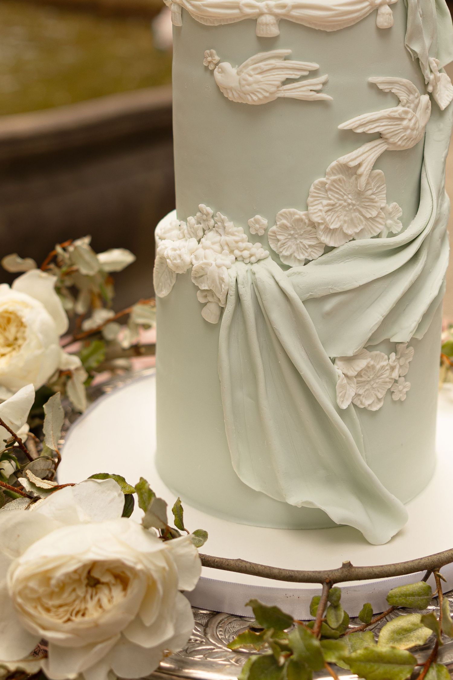 Elegant mint green wedding cake with white floral and bird fondant decorations, surrounded by cream roses on a silver stand.
