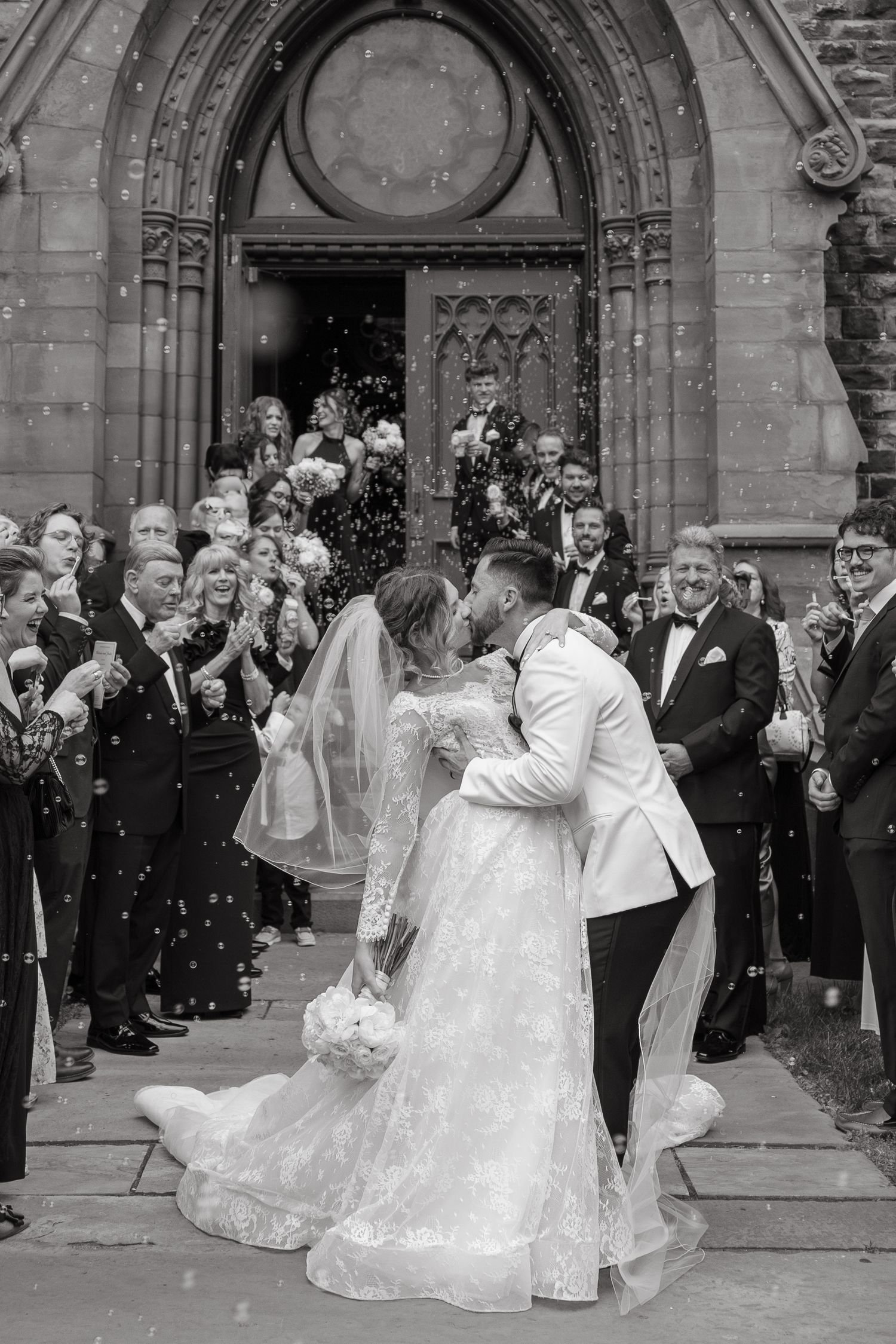 Wedding guests gather outside a Gothic church entrance as the bridal party poses in elegant white gowns.