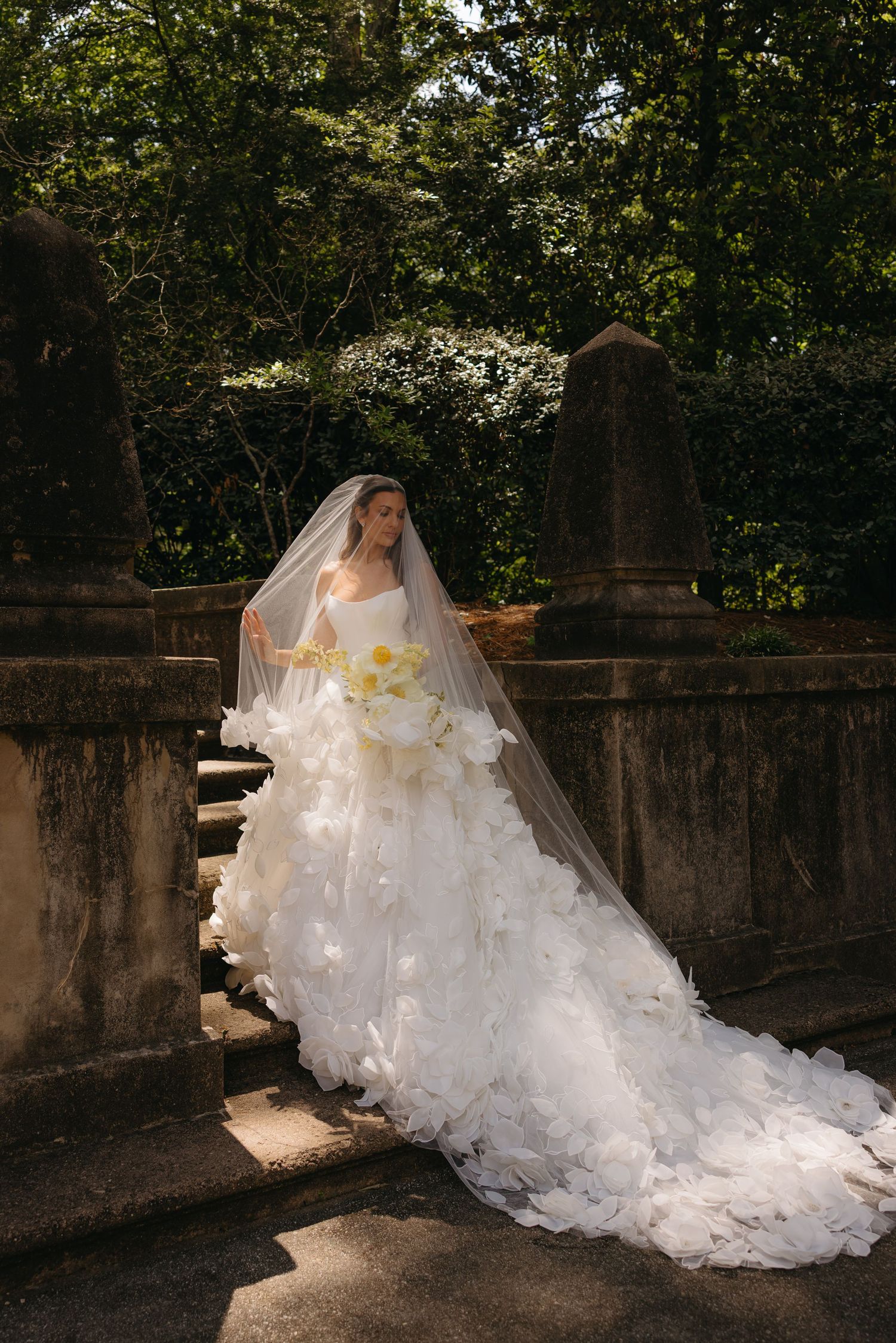 Bride in a flowing white floral ball gown with veil descends stone steps surrounded by lush greenery.