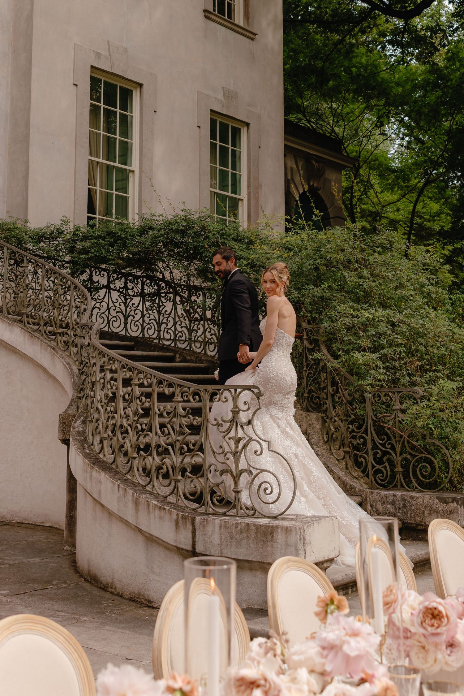 Bride and groom ascend ornate iron staircase of historic mansion with floral reception tables below.