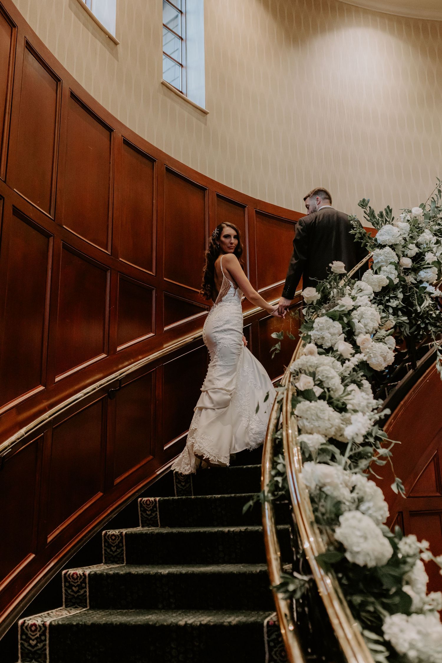 Bride and groom ascending elegant staircase decorated with white floral arrangements.