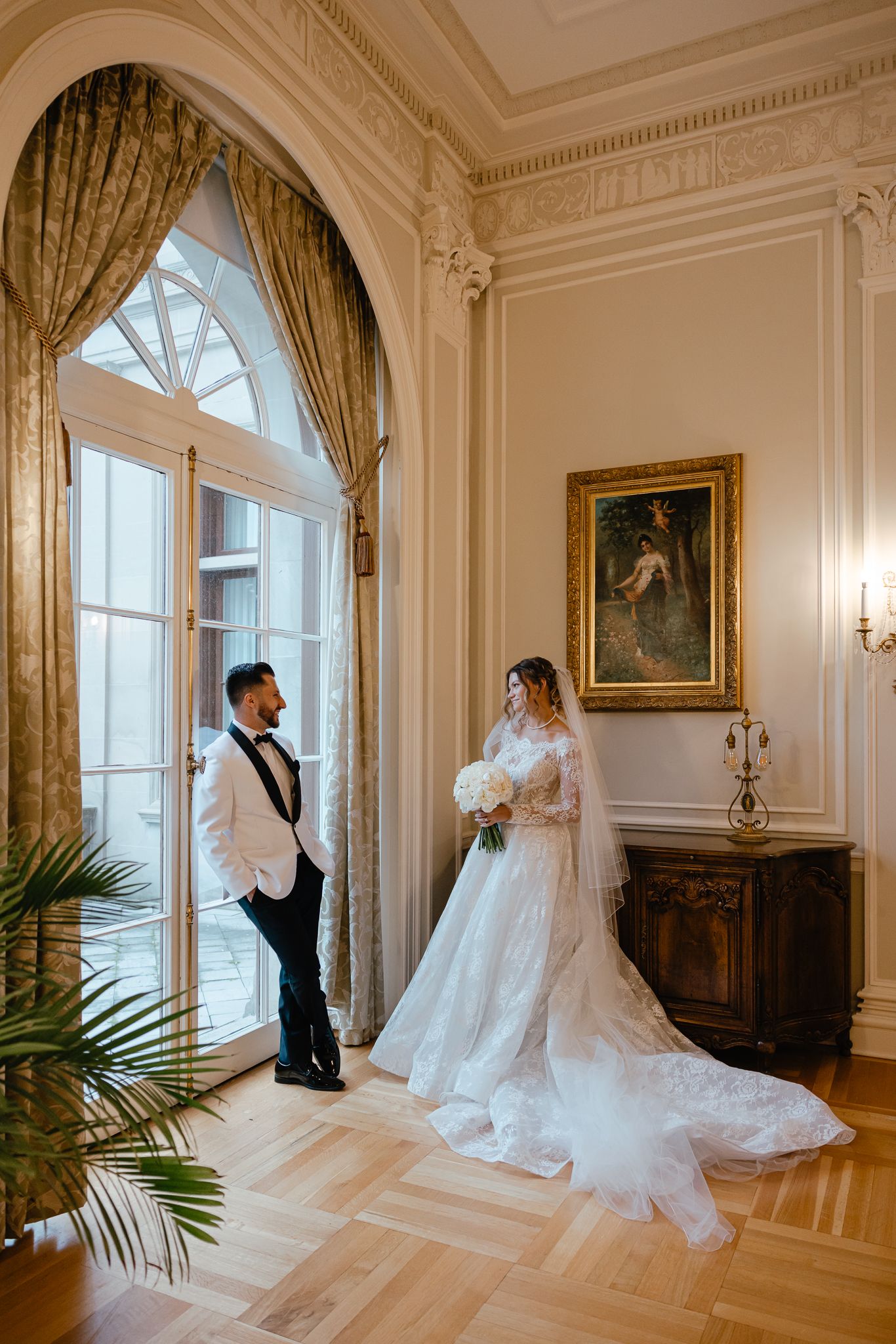 A couple shares an intimate moment in an elegant ballroom with ornate white walls and tall windows.